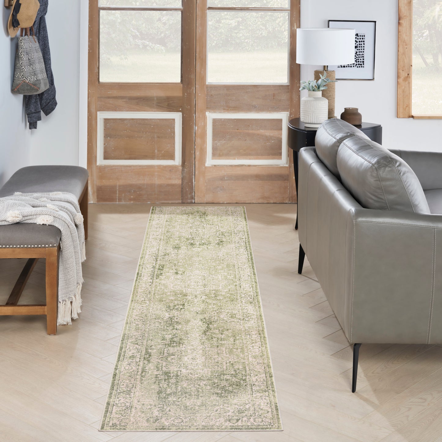 Distressed sage-green vintage runner rug with beige border along a narrow entry path between bench and gray leather sofa.