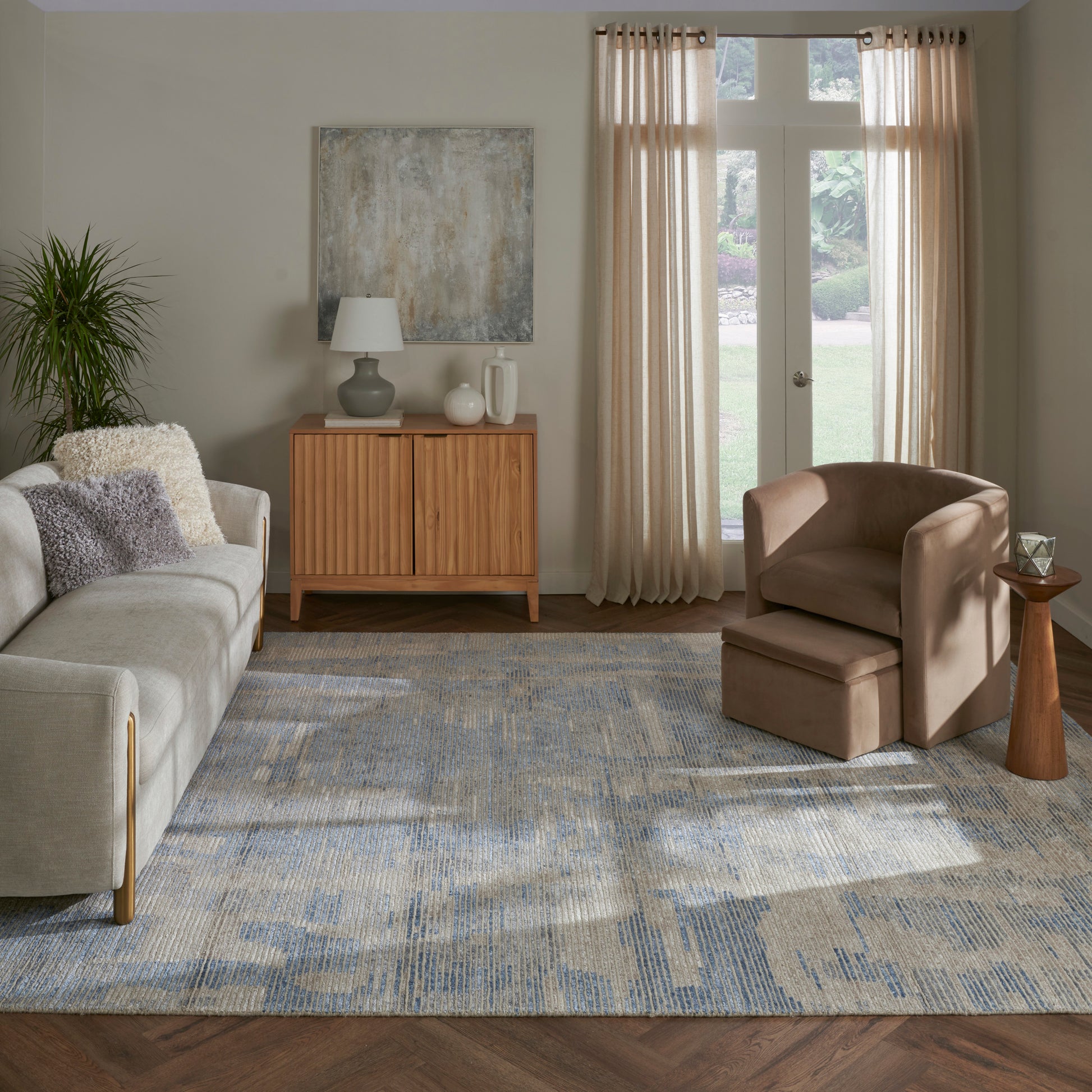 Cozy living room with light gray sofa, beige armchair & ottoman, wooden sideboard, abstract art, plant, blue-gray rug.