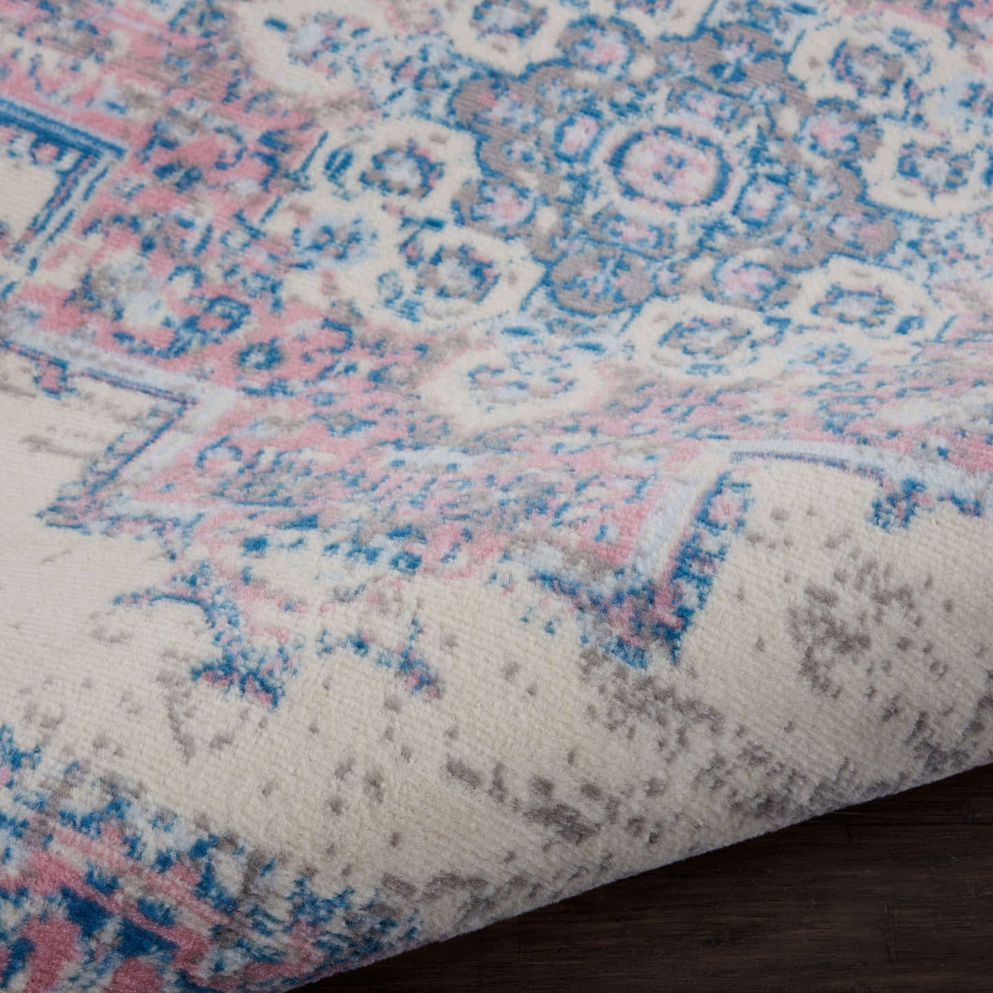 Close-up of pastel blue and pink patterned rug with cream background, showing floral motifs and a folded corner on a wooden floor.