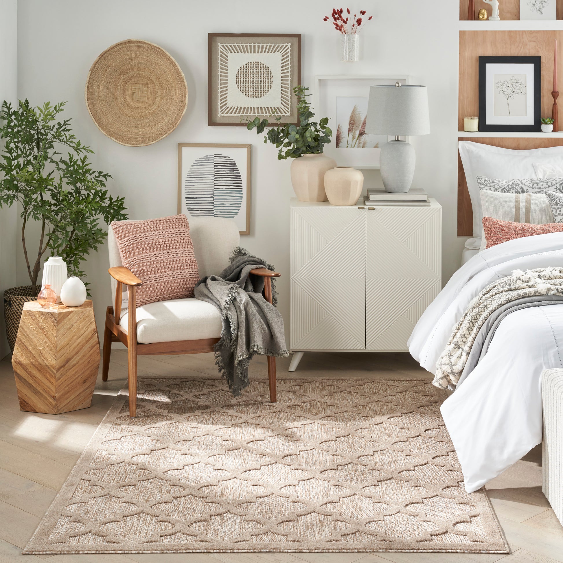 Neutral-toned bedroom with beige geometric rug, white bedding, wooden armchair, potted plant, wall art, and ceramic vases.