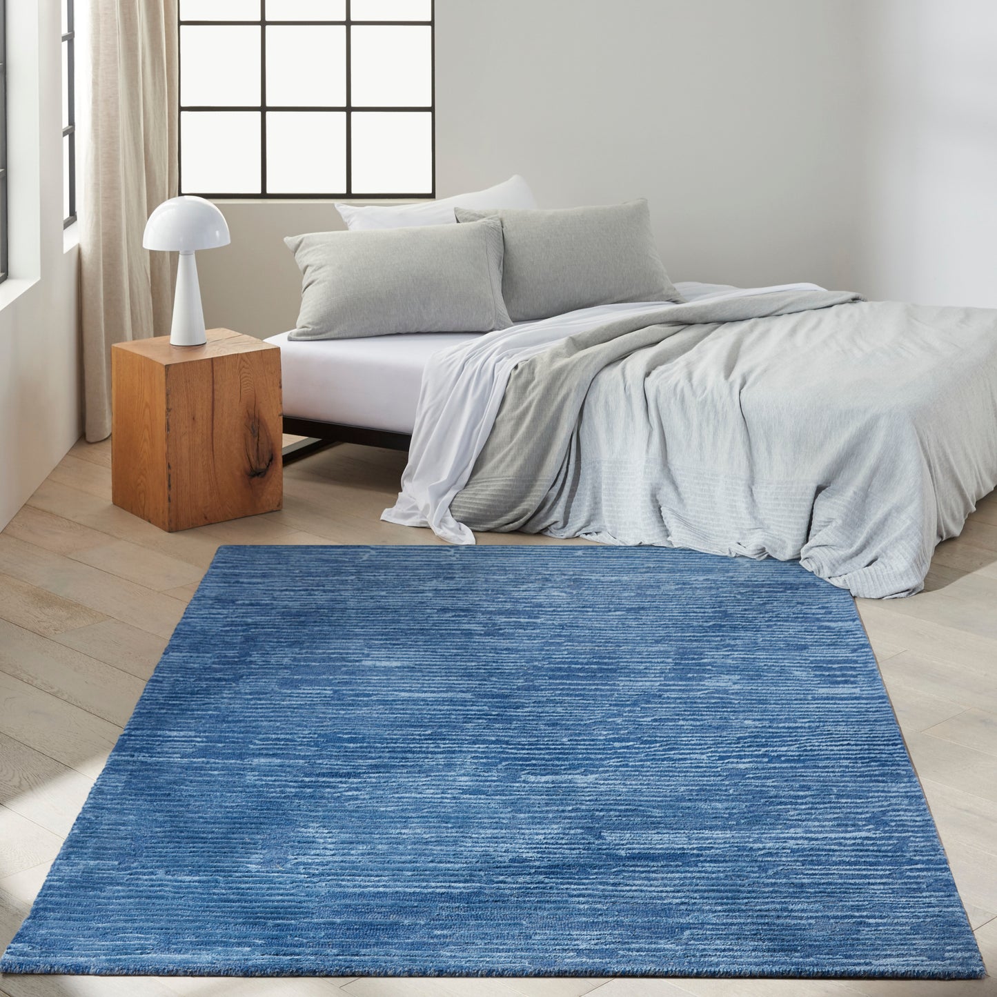 Blue rectangular flat-weave area rug for a modern bedroom with gray bedding and wooden nightstand.