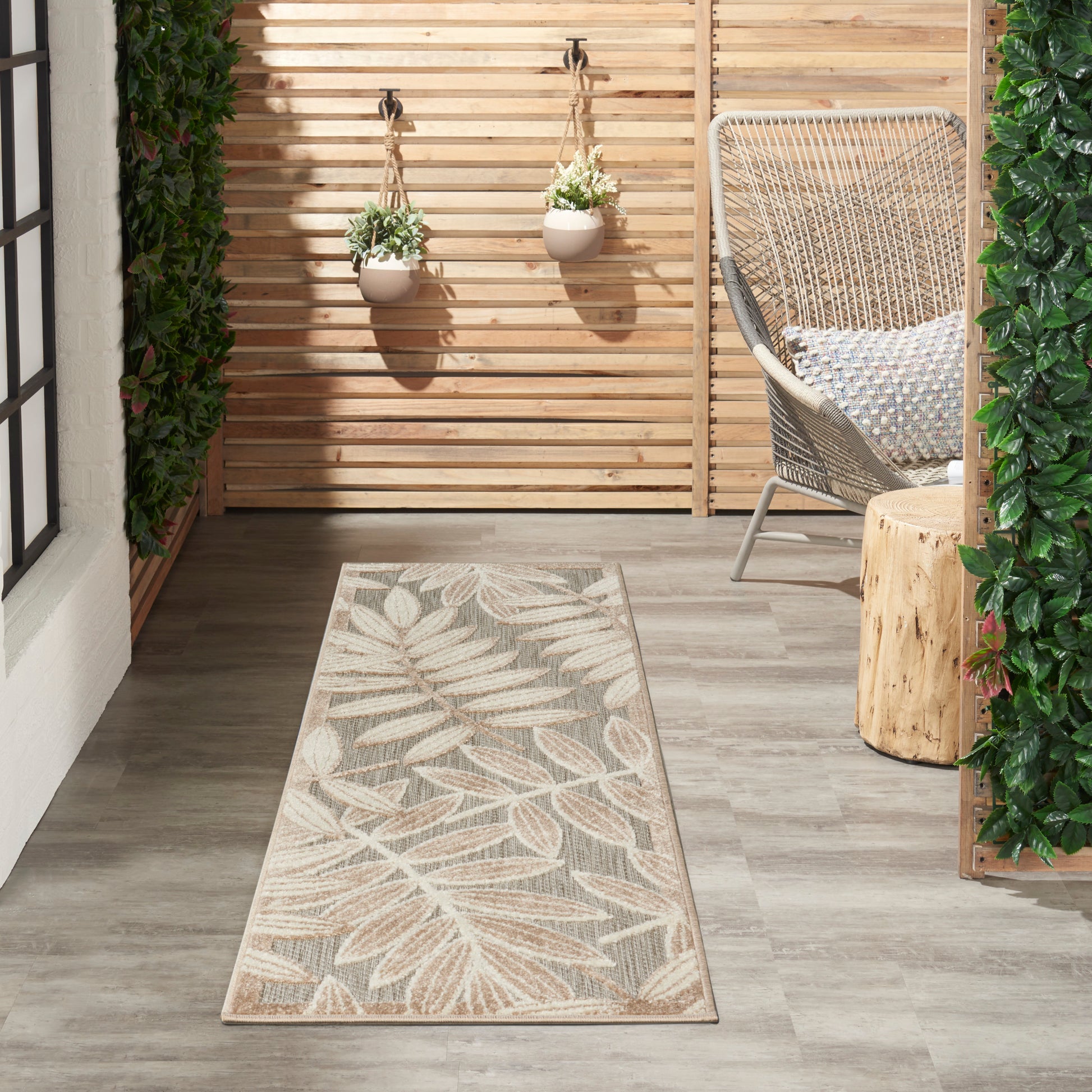Outdoor patio runner rug with beige leaf pattern on gray floor, beside wicker chair, hanging planters and wood stump table.
