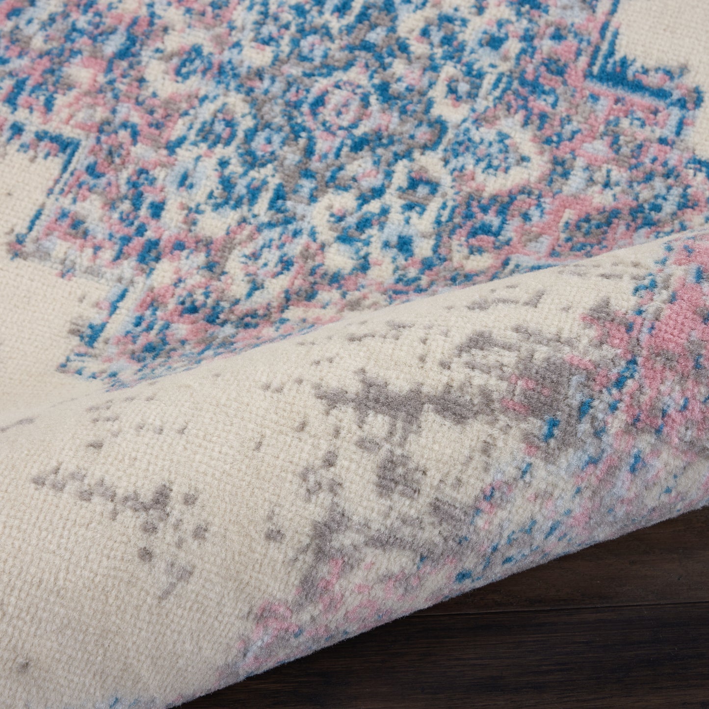 Beige woven oriental rug with blue and pink floral pattern, folded corner on dark wood floor.