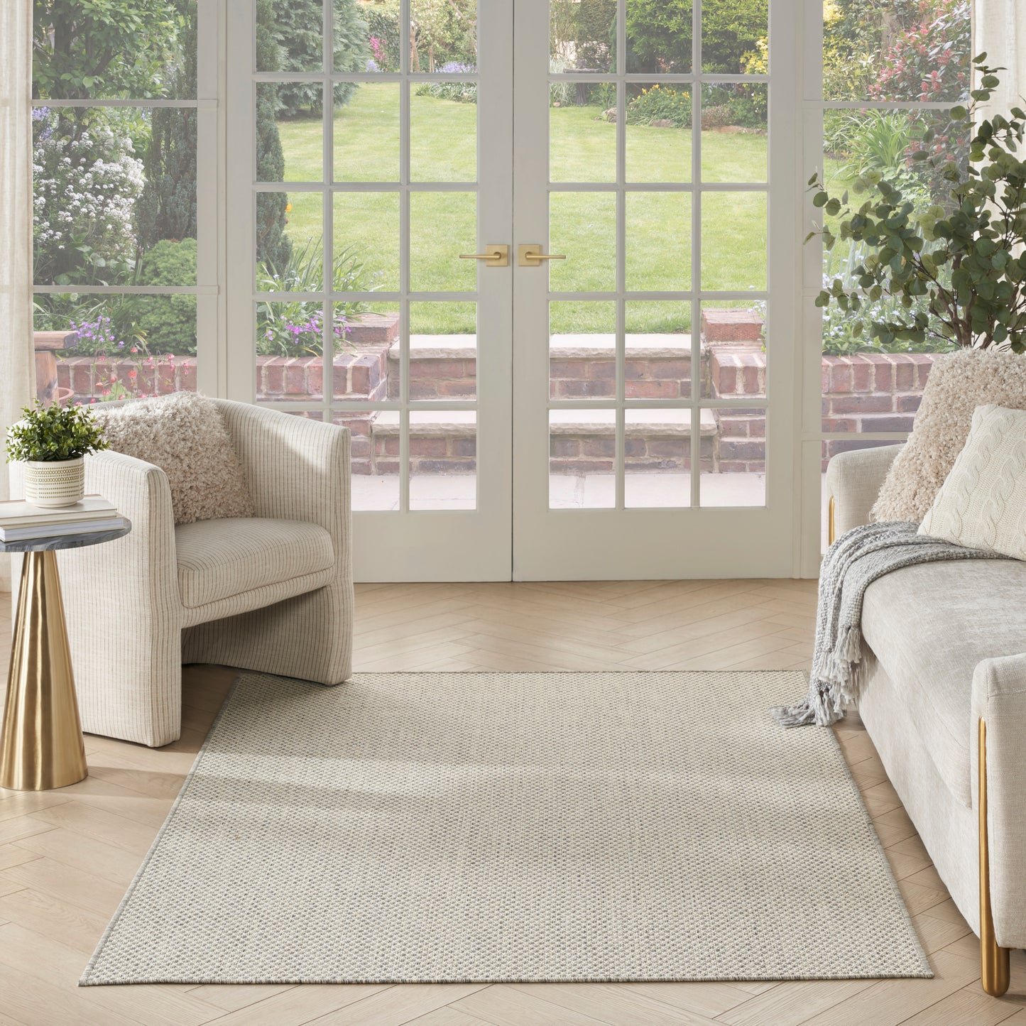 Bright living room with cream sofas, beige textured rug, glass French doors, gold accents, leafy plants, neutral cozy decor.