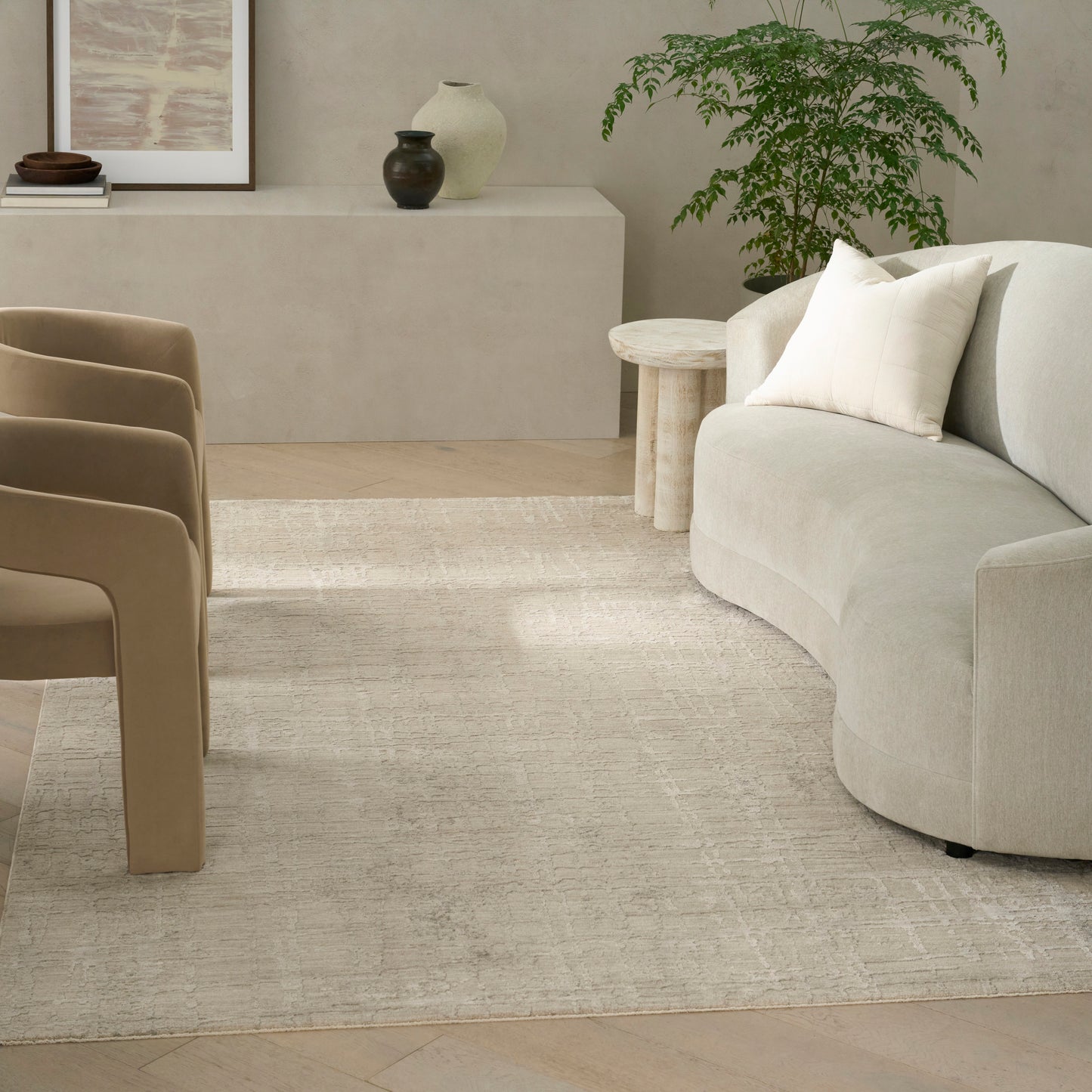 Beige modern area rug (8' x 10') in a living room scene with a curved beige sectional, tan chairs, a round wooden side table and a potted plant.