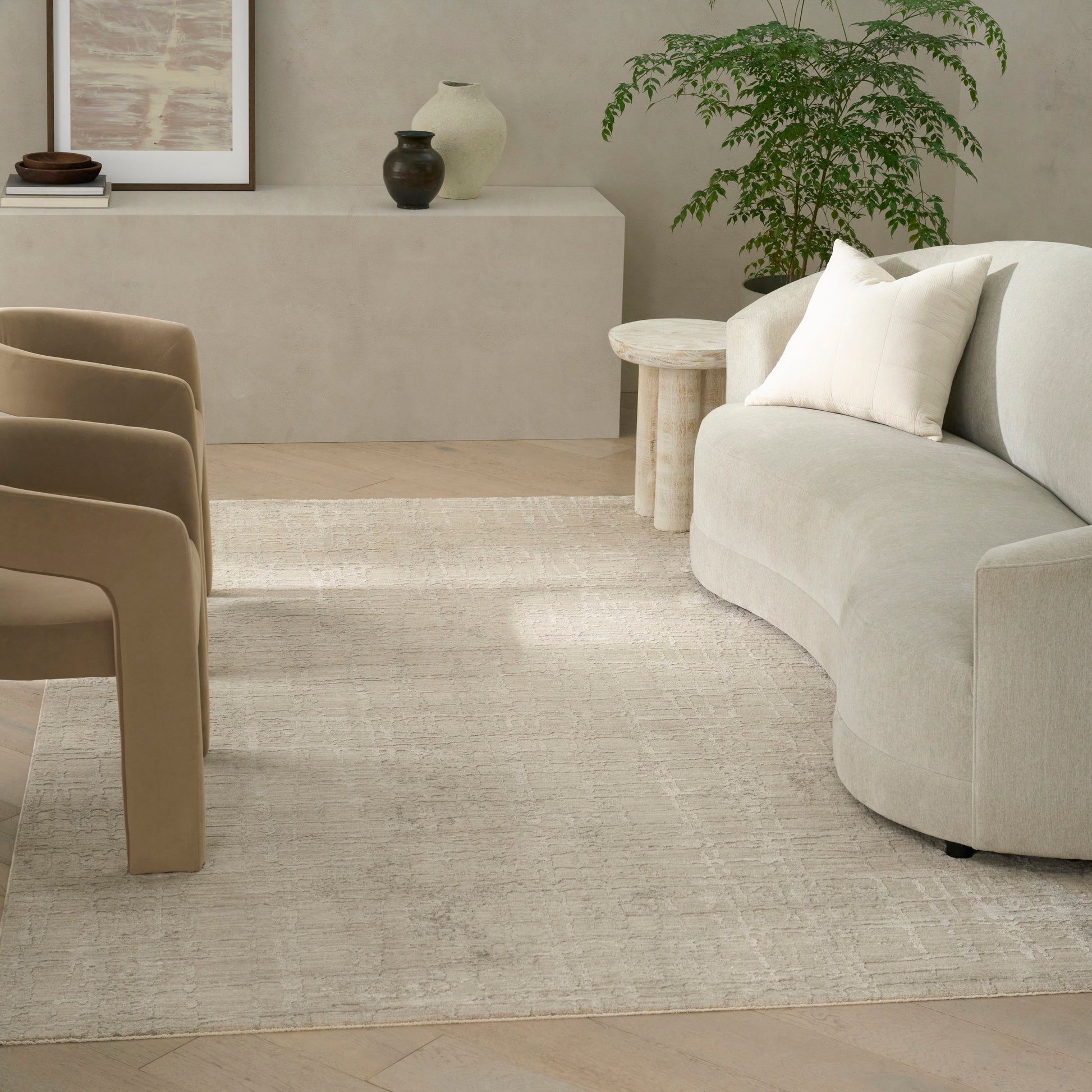 Beige modern area rug (8' x 10') in a living room scene with a curved beige sectional, tan chairs, a round wooden side table and a potted plant.