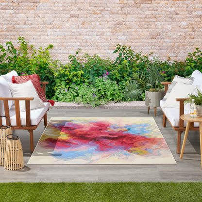 Outdoor patio with a square abstract area rug in red, blue & yellow on beige, between wooden-framed white-cushion chairs.