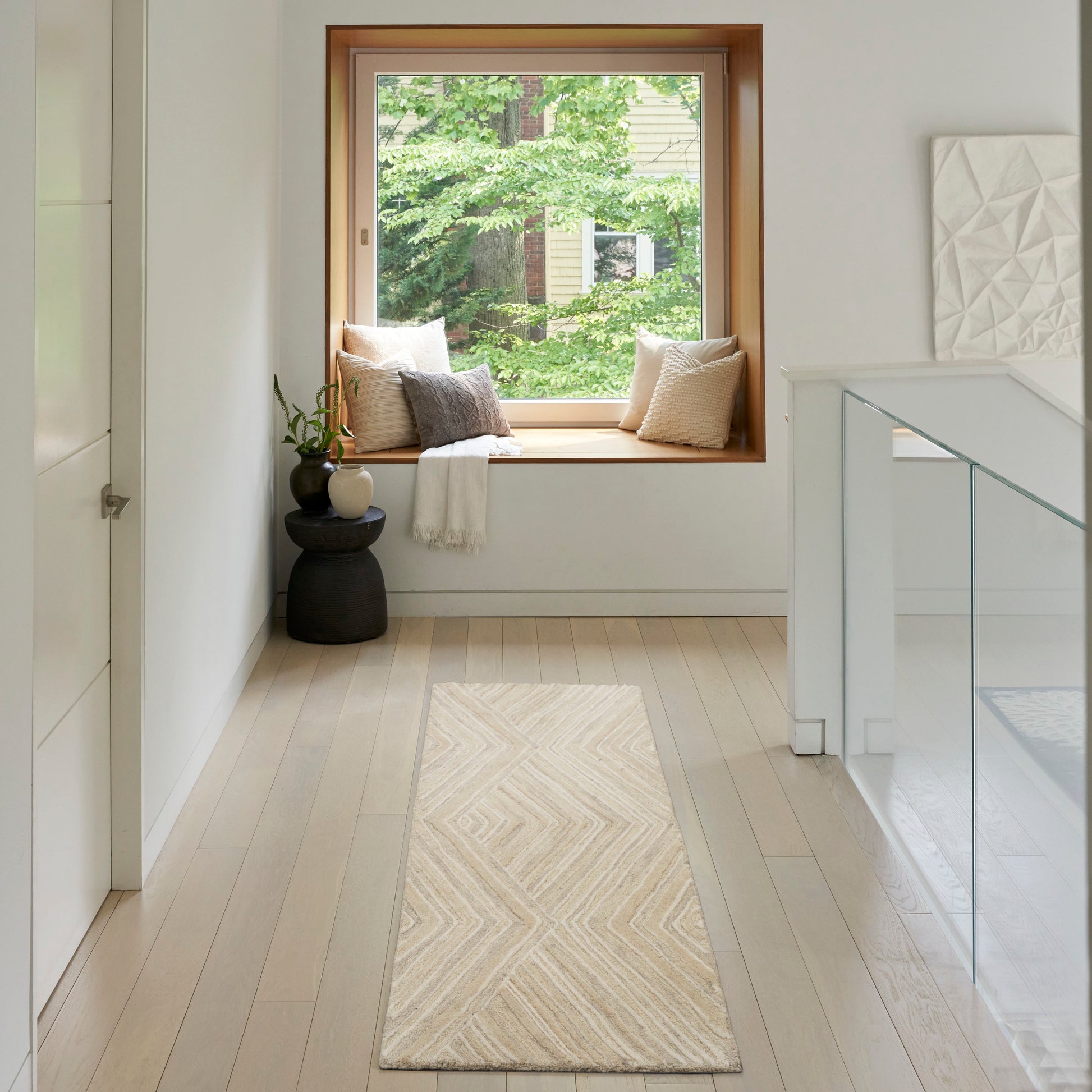 Bright hallway with a wooden-framed window seat, beige cushions, cream rug, white walls, glass railing; greenery outside