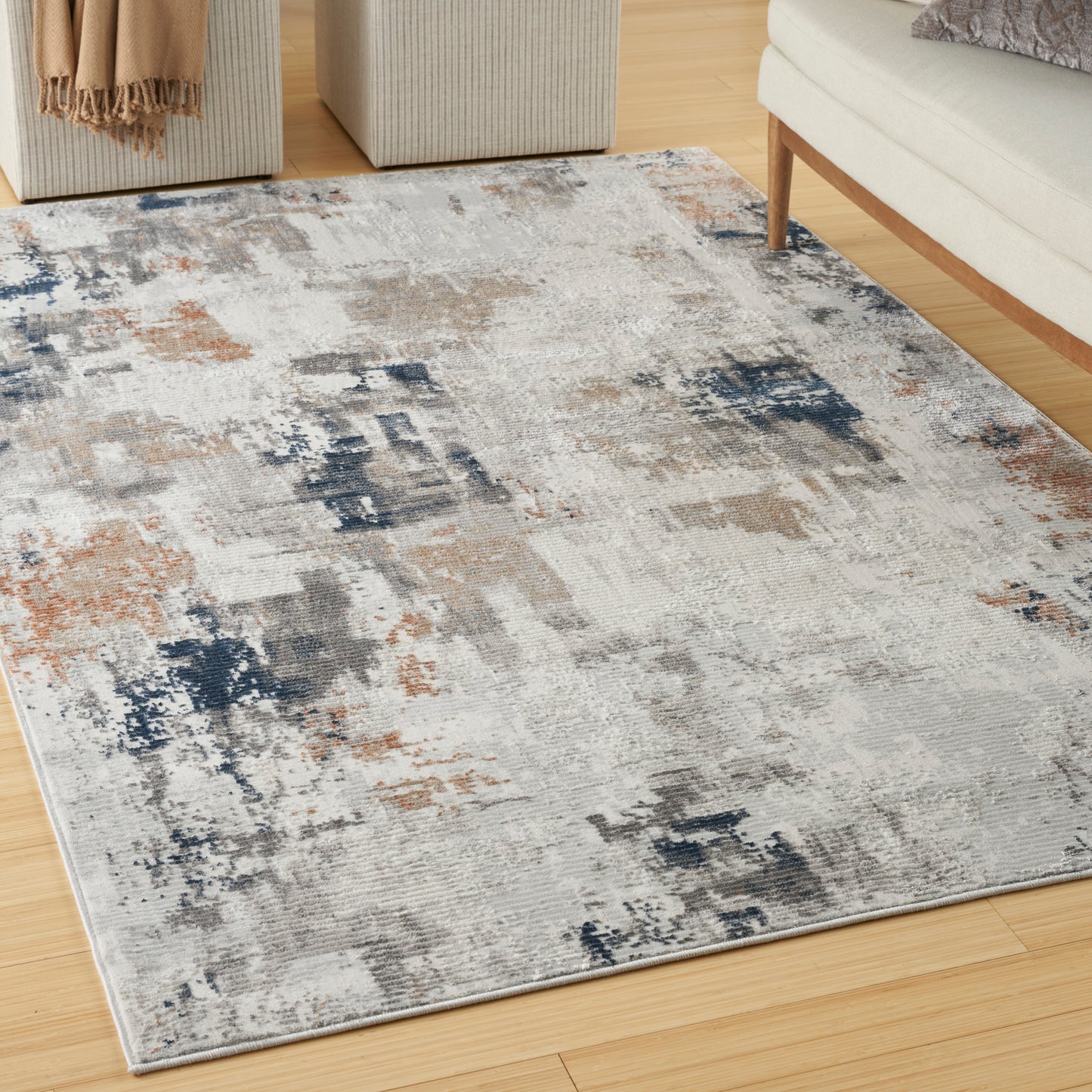 Rectangular abstract-pattern area rug in gray, navy & tan, soft pile on light wood floor; complements beige ottomans & sofa.