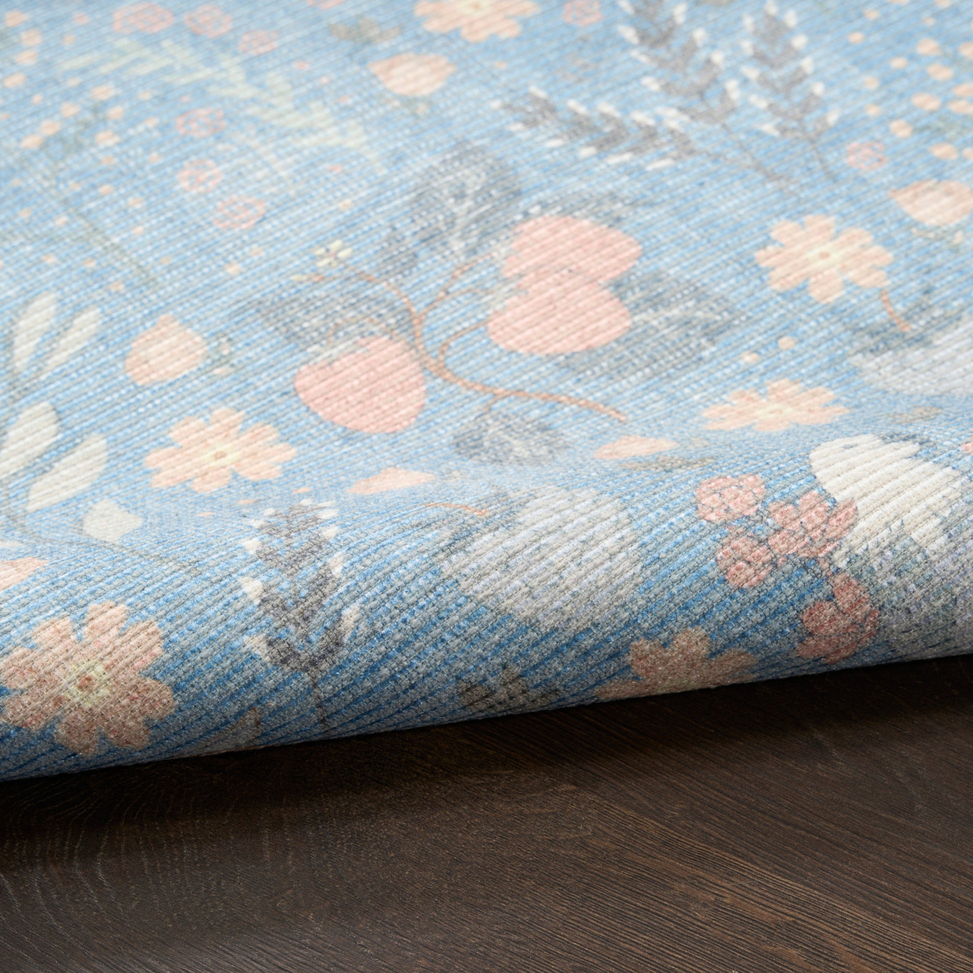 Blue floral woven rug with pale pink flowers and gray leaves, textured weave, folded edge on dark wood surface.