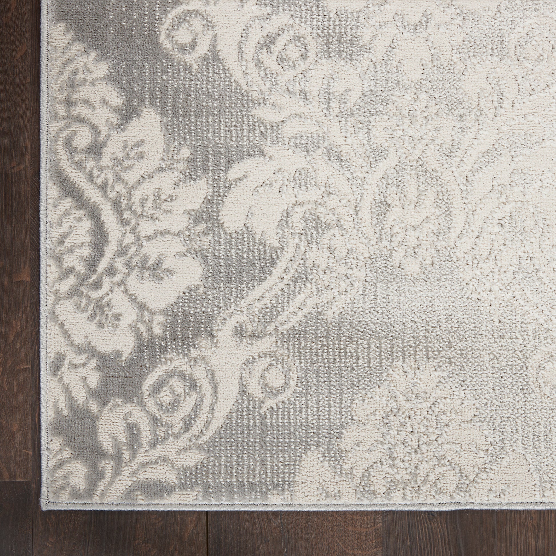 Rectangular woven rug with ivory damask floral pattern on gray background, on dark wood floor.