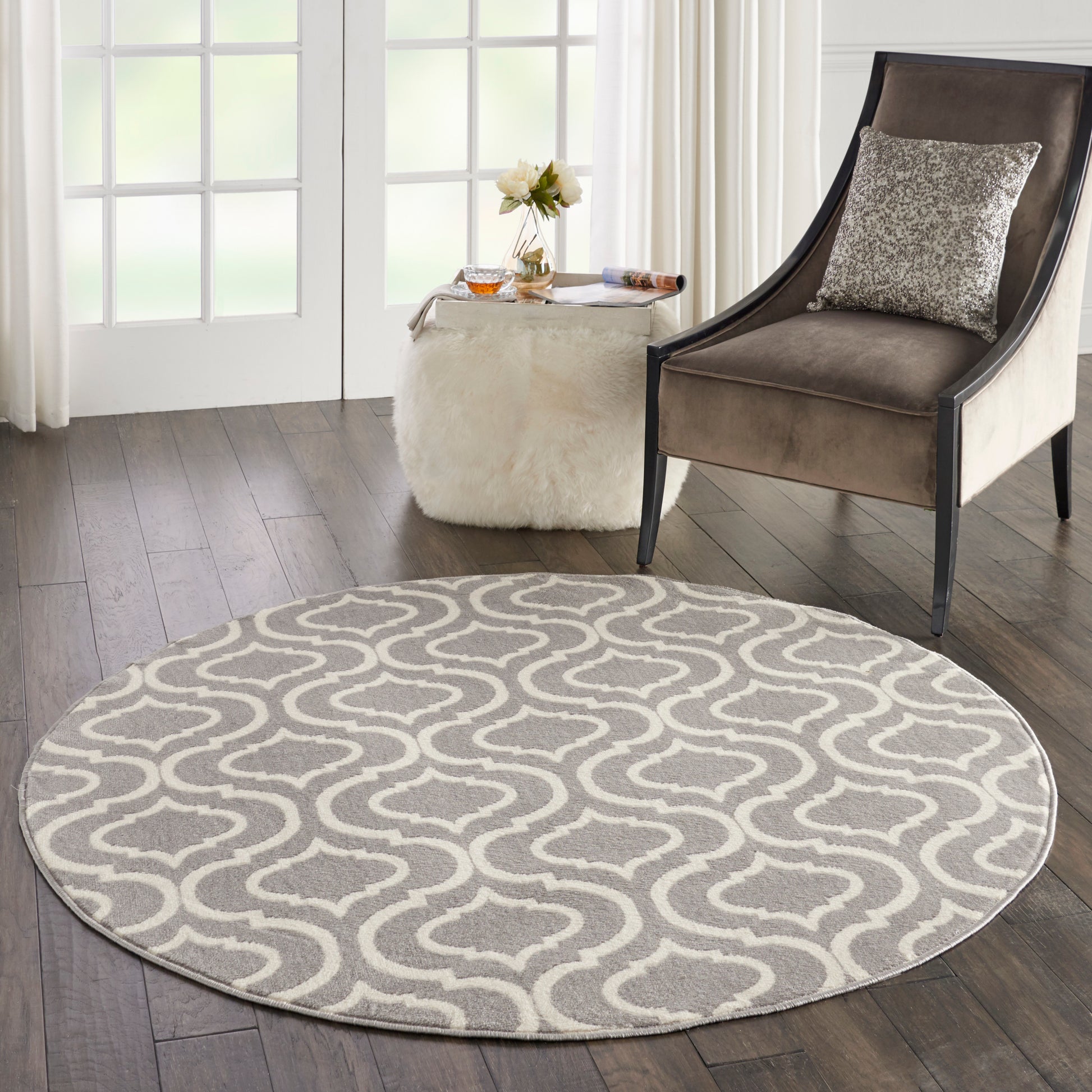 Round gray rug with white wavy lattice pattern on dark wood floor, beside a brown upholstered armchair and white fur ottoman.