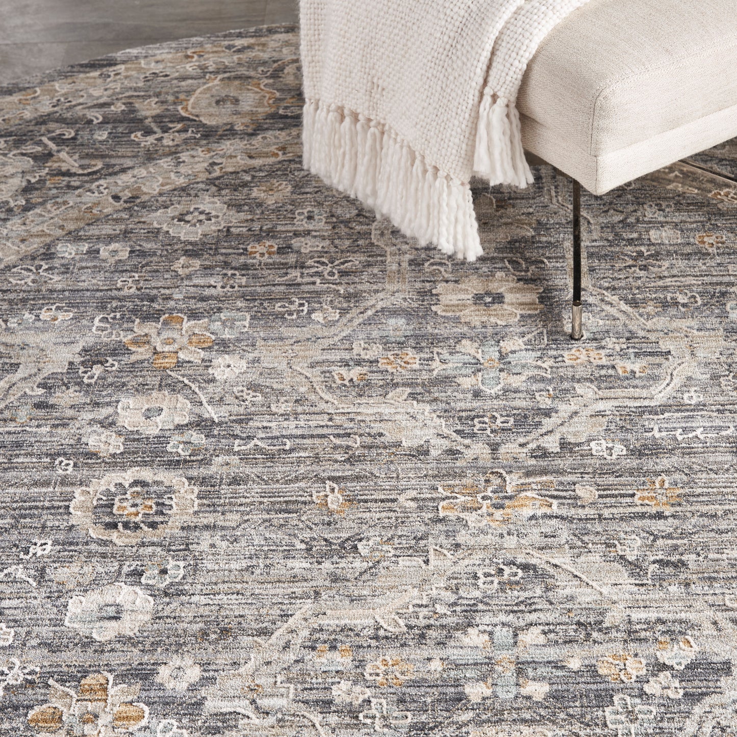 Muted floral grey area rug with ivory and pale blue accents under a beige sofa and cream fringed throw.