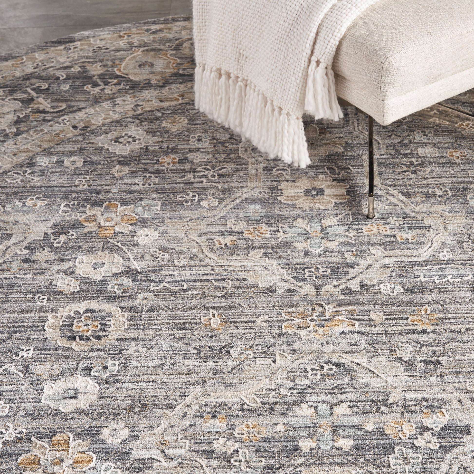 Muted floral grey area rug with ivory and pale blue accents under a beige sofa and cream fringed throw.