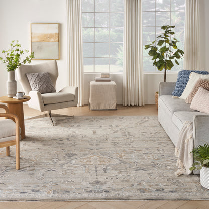 Bright neutral living room with grey sofa, beige armchairs, woven rug, light wood tables, large windows, and potted plants.