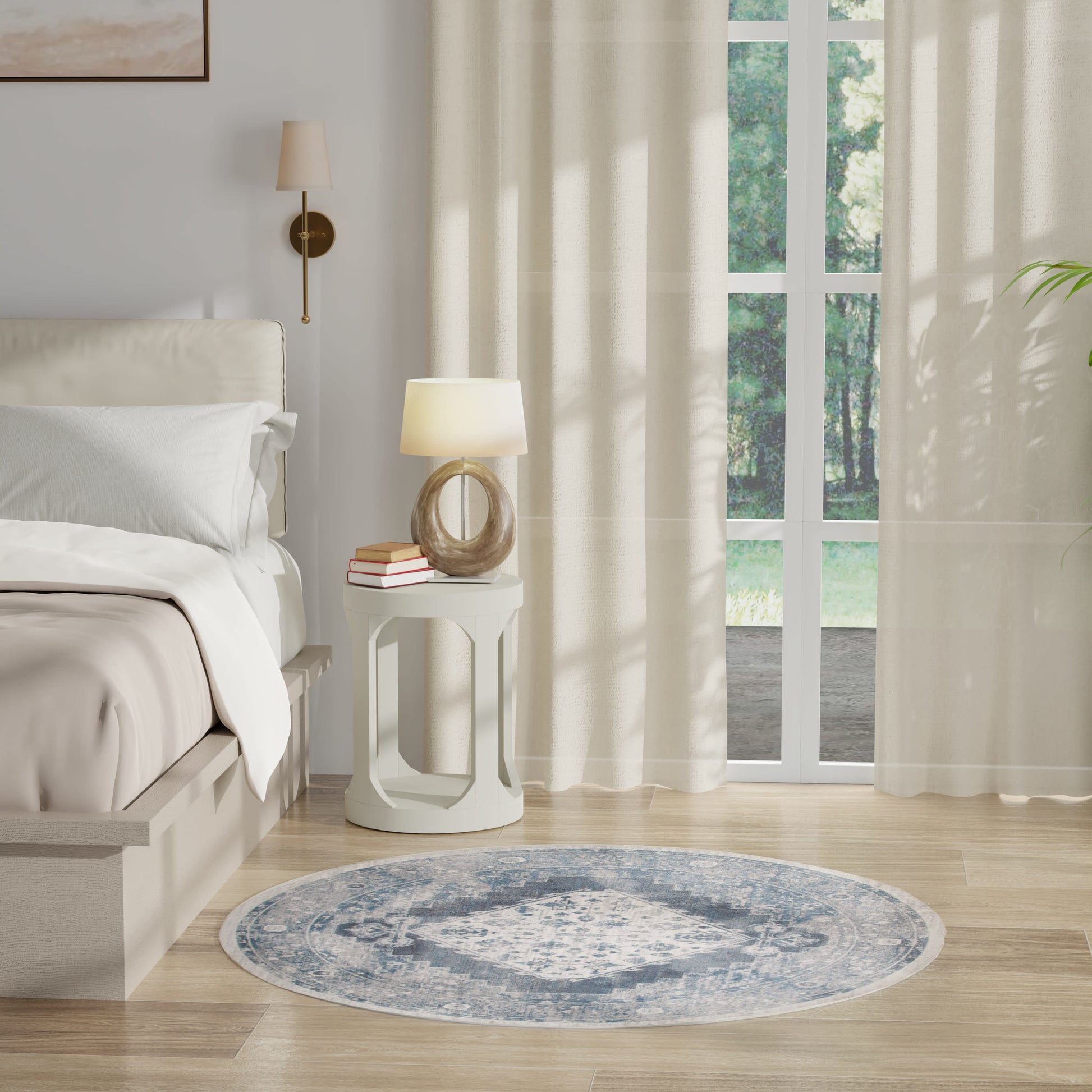 Round blue patterned rug on light wood floor beside white round nightstand, ring lamp and books; beige bed with linens.