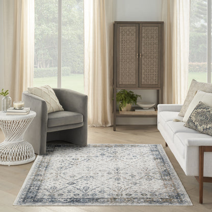 Blue-gray patterned area rug on light wood floor in a living room with gray armchair, white sofa, cream curtains, and wicker cabinet.
