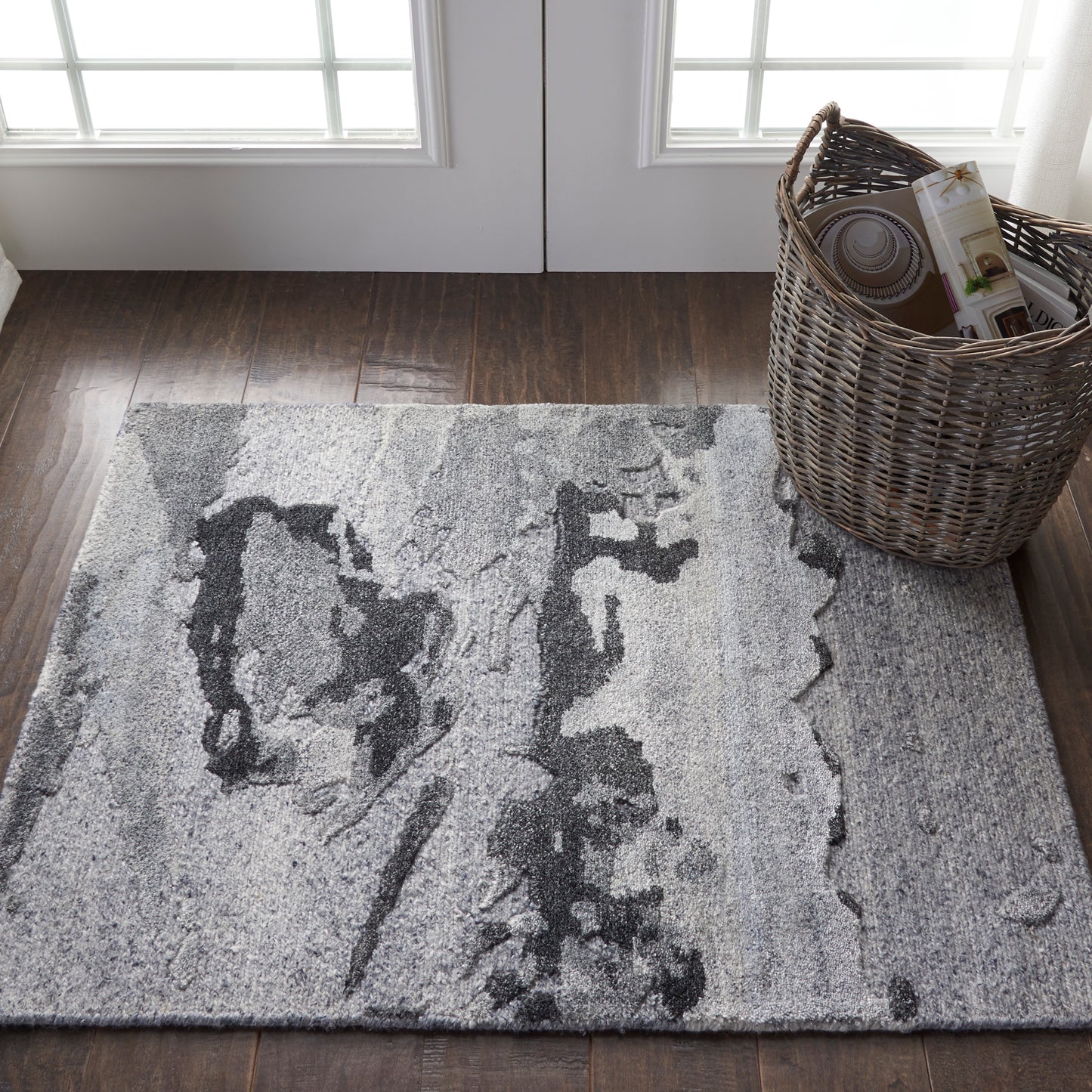 Gray abstract-pattern area rug on dark wood floor near glass-paneled doors, with a wicker basket of magazines.