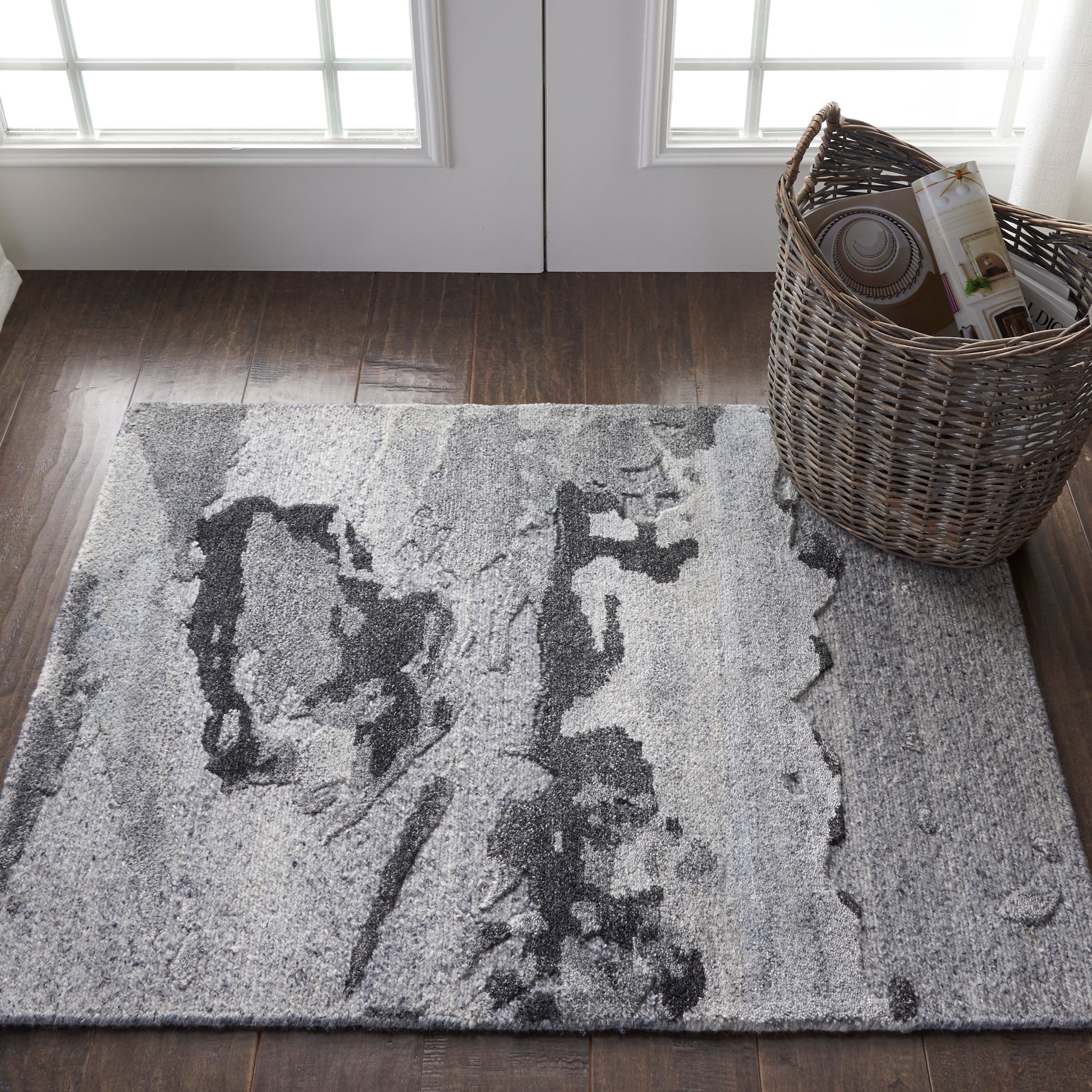 Gray abstract-pattern area rug on dark wood floor near glass-paneled doors, with a wicker basket of magazines.