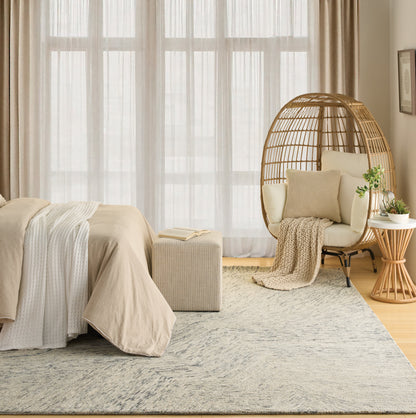 Neutral beige rug in a bedroom scene with a bed in taupe linens, a wicker egg chair, and a small wood side table by sheer-curtained windows.