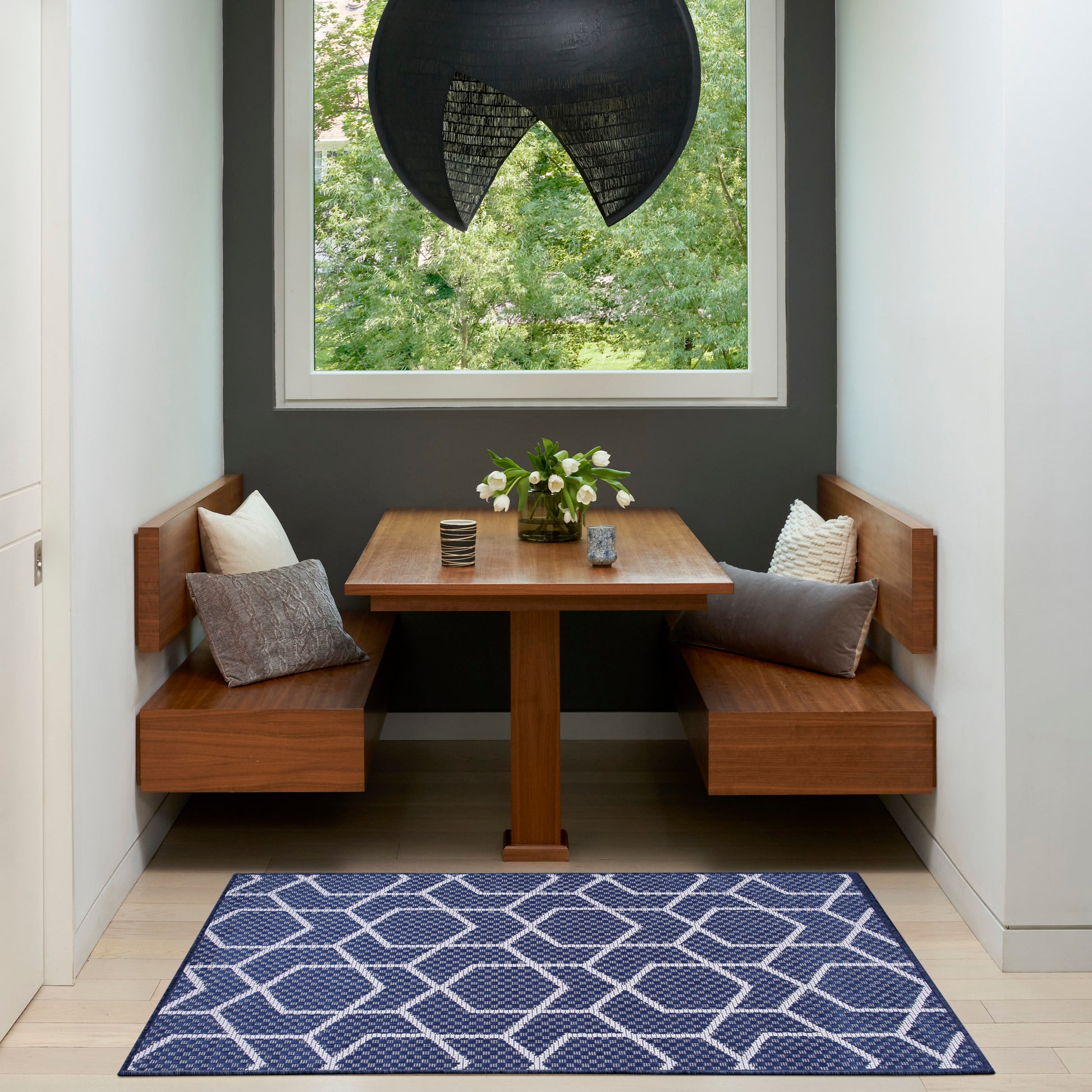 Wooden dining nook with benches, gray cushions, vase of white flowers, table, black pendant light, and blue geometric rug.