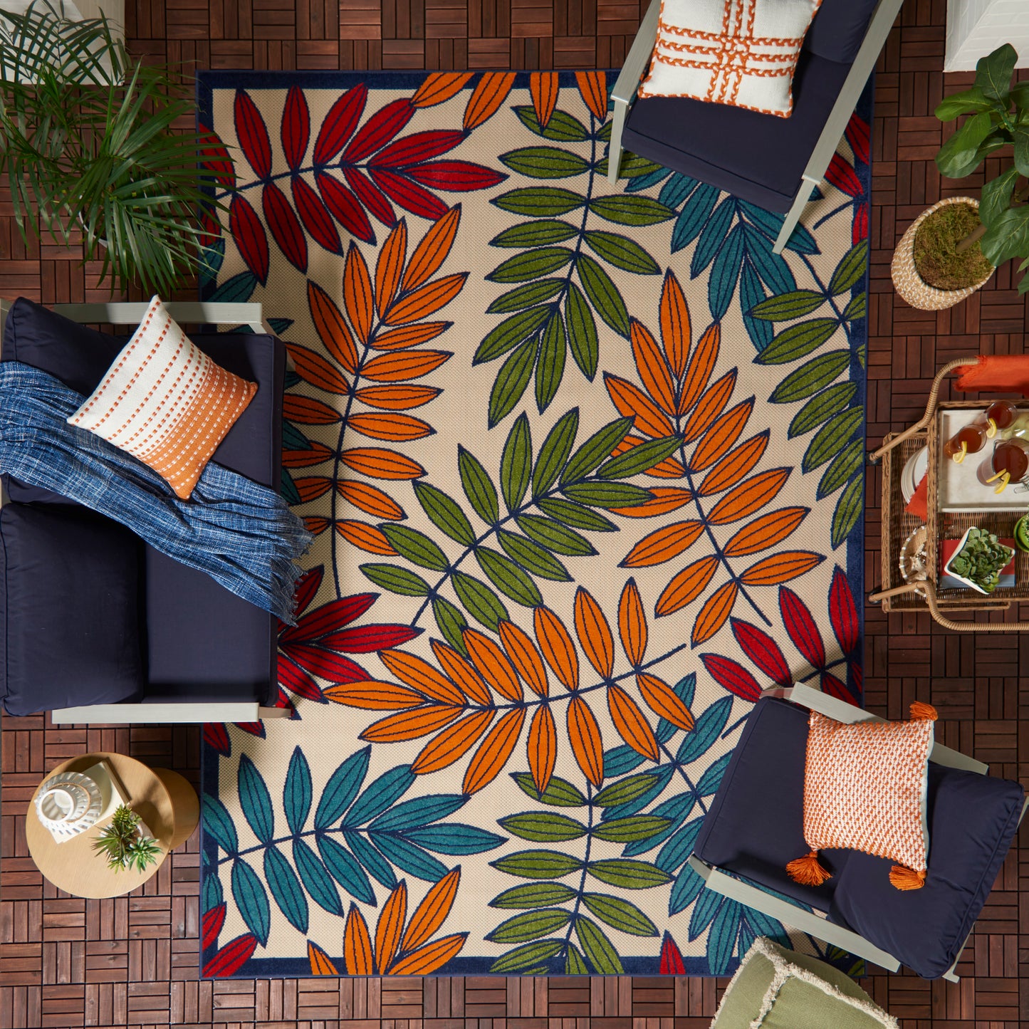 Top-down view of an 8' x 11' tropical leaf rug in orange, red, blue, green and teal, surrounded by navy sofas, throws, pillows and plants.