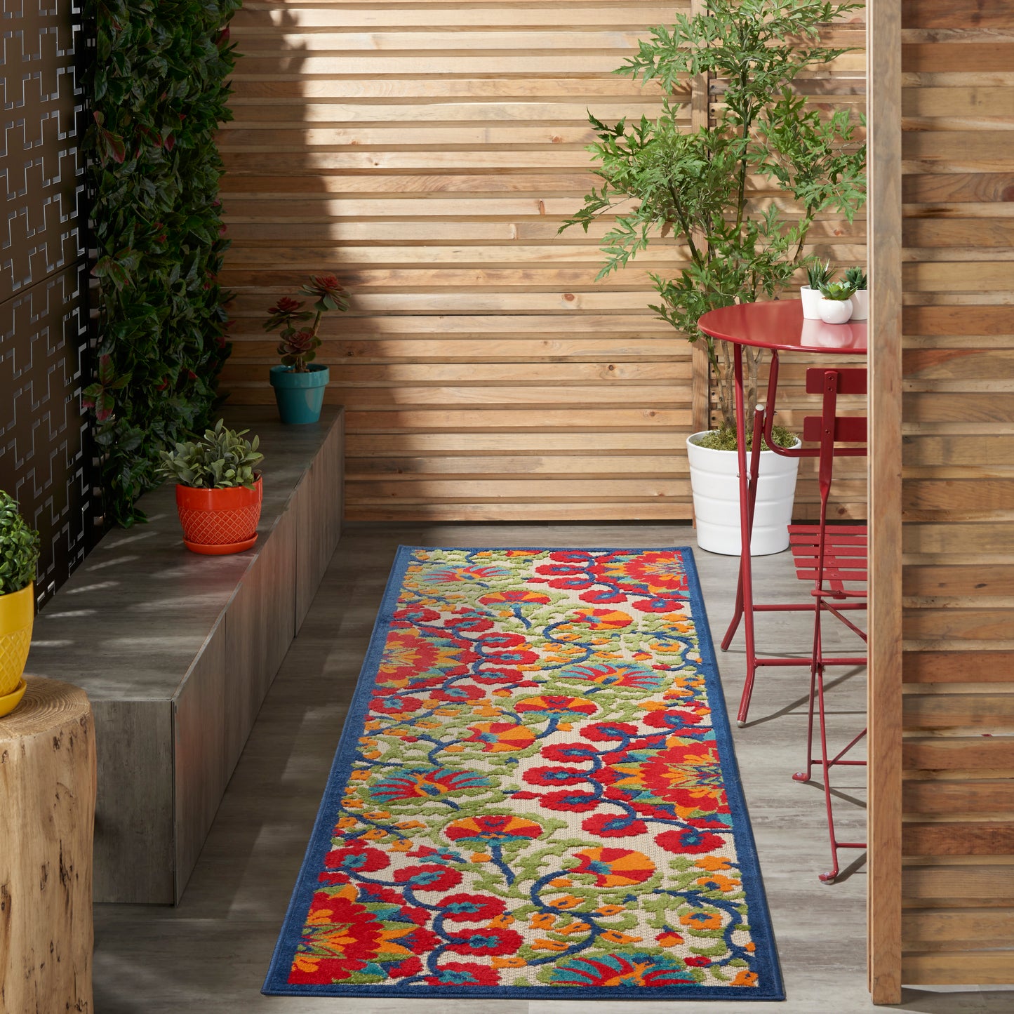 Outdoor runner rug with blue border and bright multicolor floral print on a wooden patio beside a red bistro table.