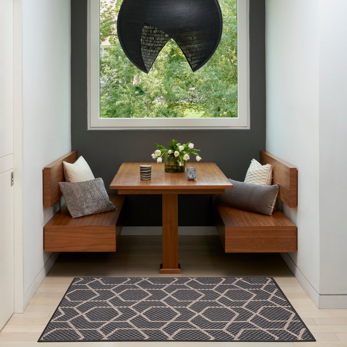Wood dining nook with built-in benches, gray cushions, square table, black woven pendant, vase of flowers, geometric rug.