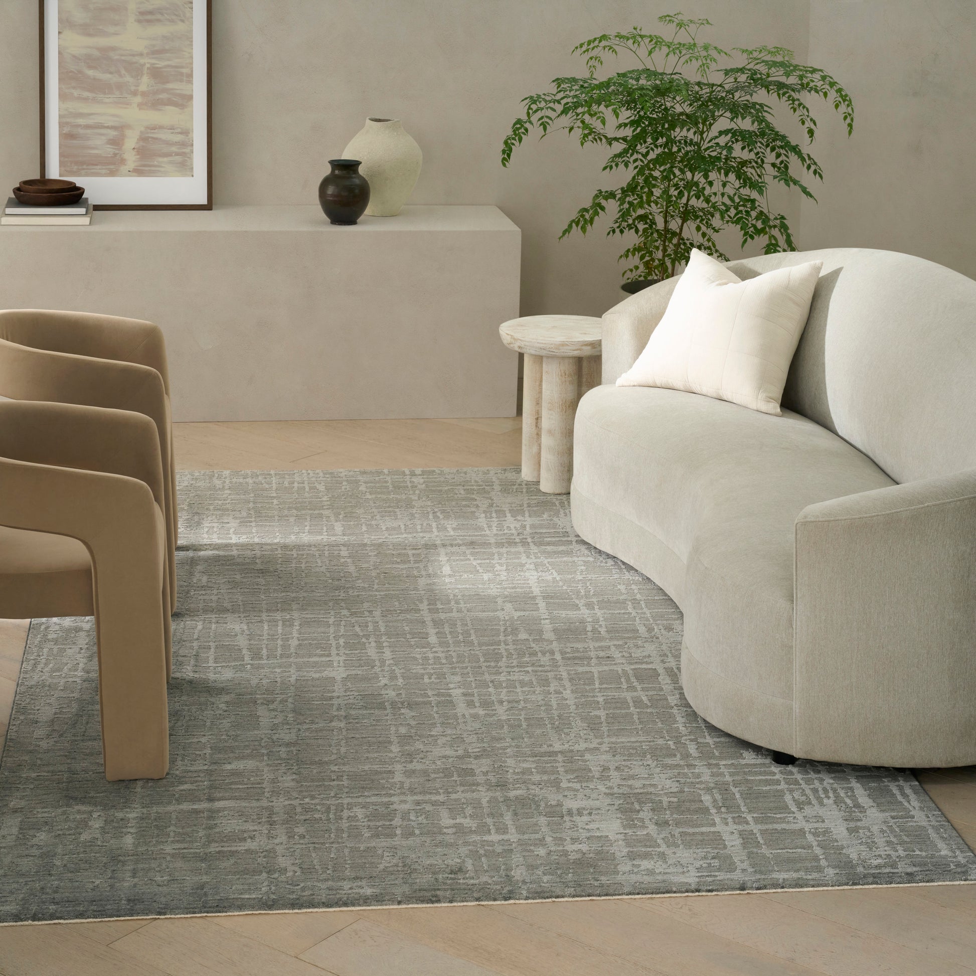 Neutral living room with curved beige sofa, white pillow, tan chairs, round side table, light gray patterned rug, greenery.