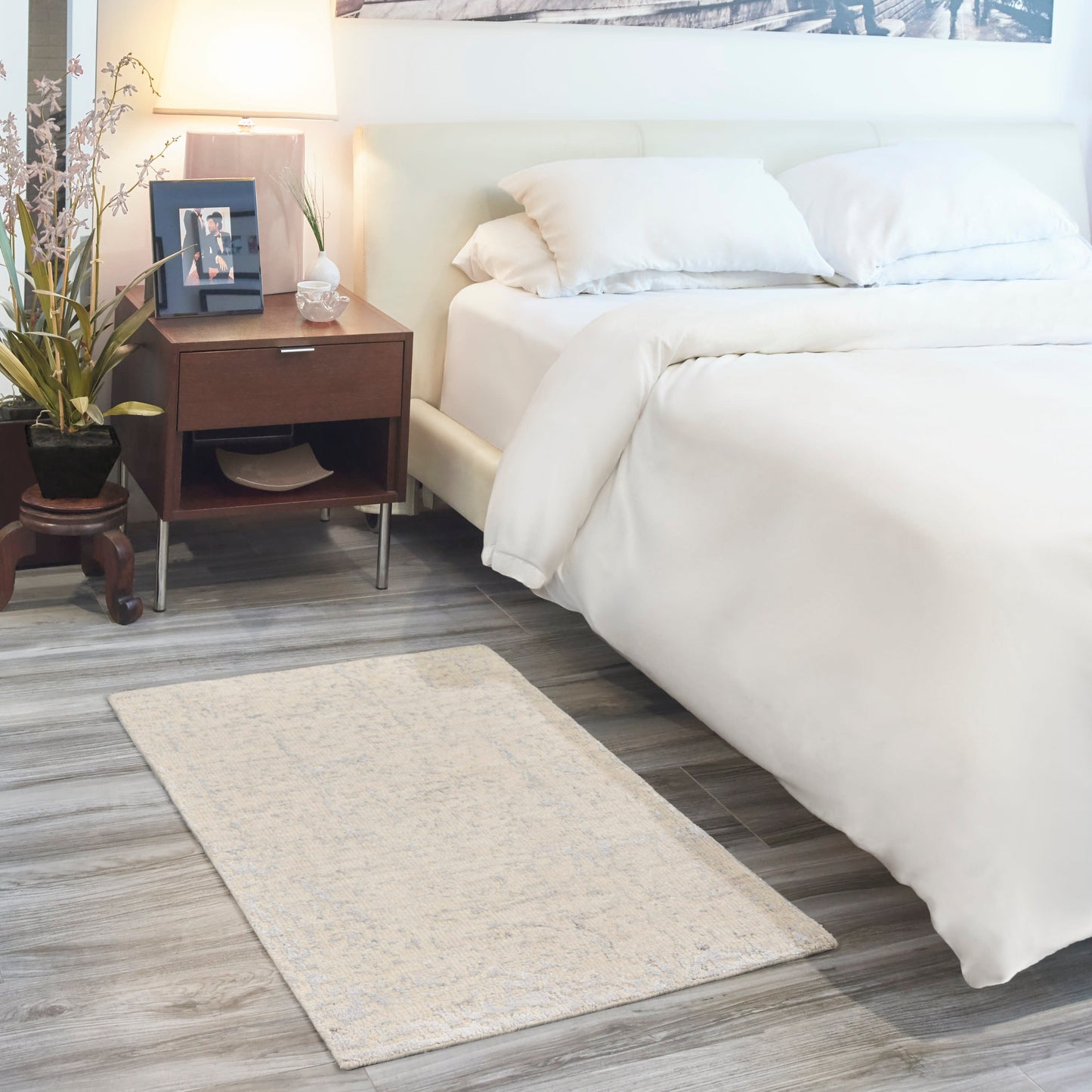 White bed with plush bedding beside dark wood nightstand, table lamp, plant, framed photo, & beige patterned area rug.