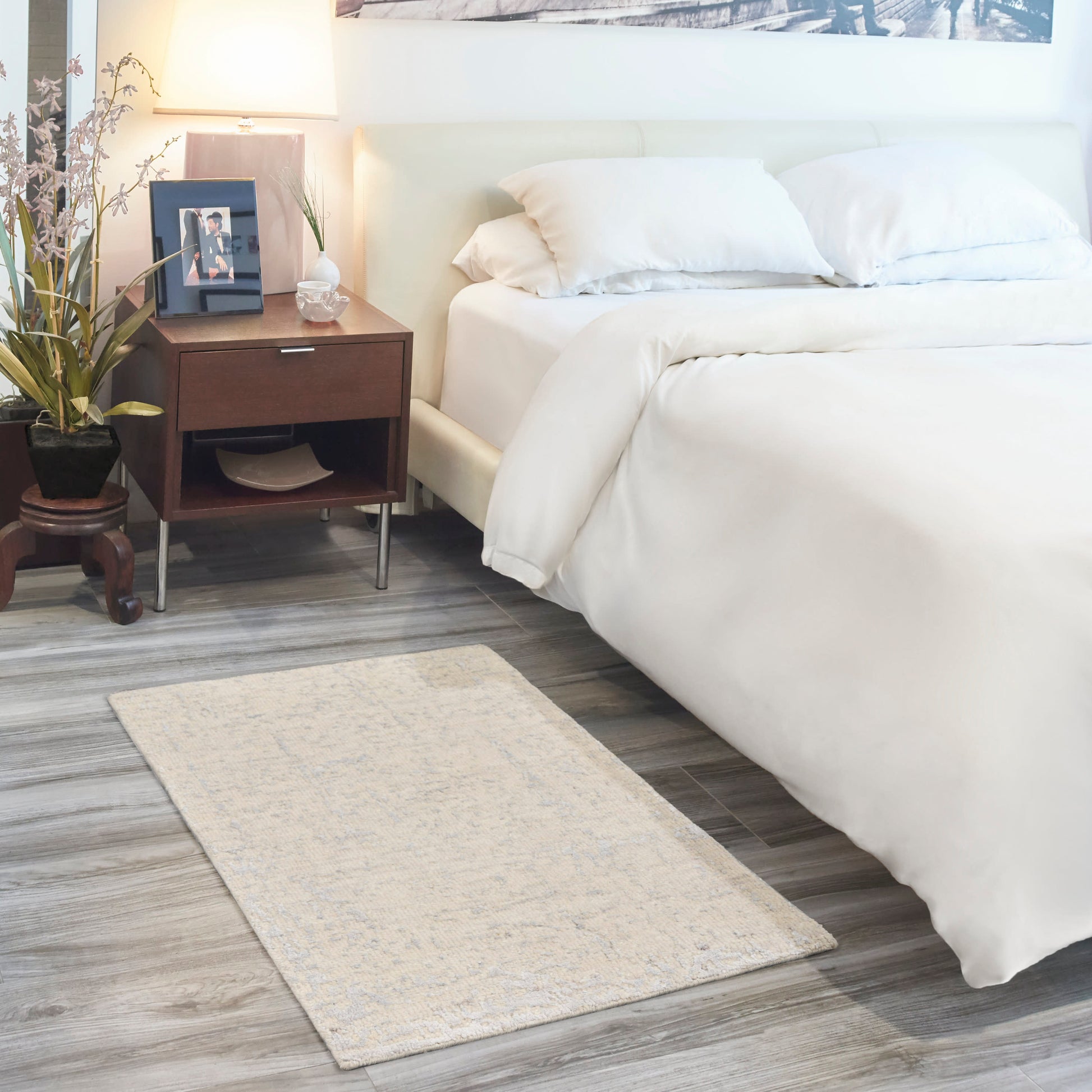 White bed with plush bedding beside dark wood nightstand, table lamp, plant, framed photo, & beige patterned area rug.