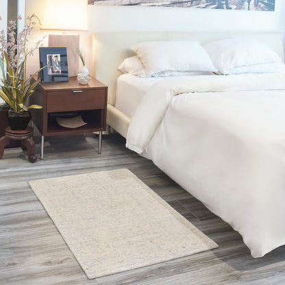 White bed with plush bedding beside dark wood nightstand, table lamp, plant, framed photo, & beige patterned area rug.