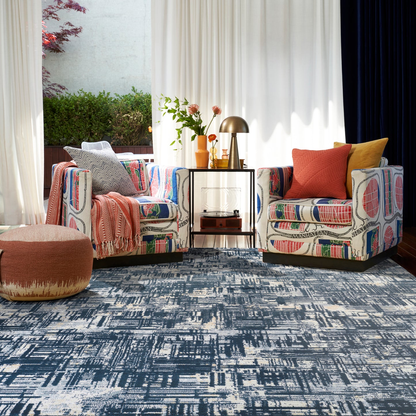 Two bold patterned armchairs with orange and mustard cushions, a round ottoman, glass side table, brass lamp, and blue rug.