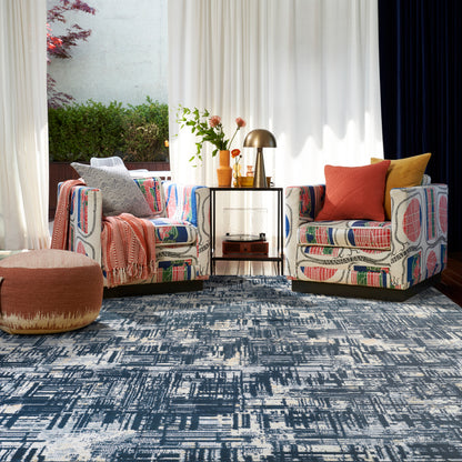 Two bold patterned armchairs with orange and mustard cushions, a round ottoman, glass side table, brass lamp, and blue rug.
