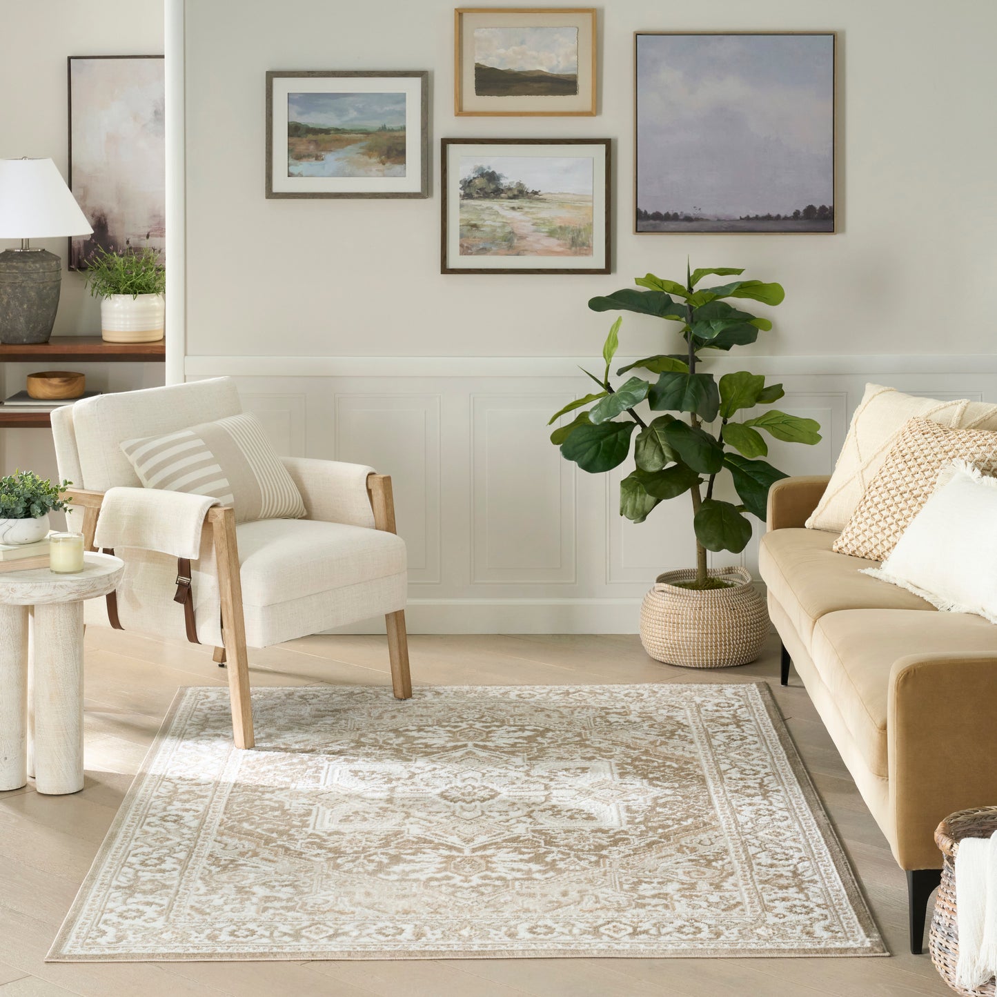 Cozy neutral living room with beige sofa, light wood armchair, ornate beige rug, fiddle-leaf fig, and framed landscape art.