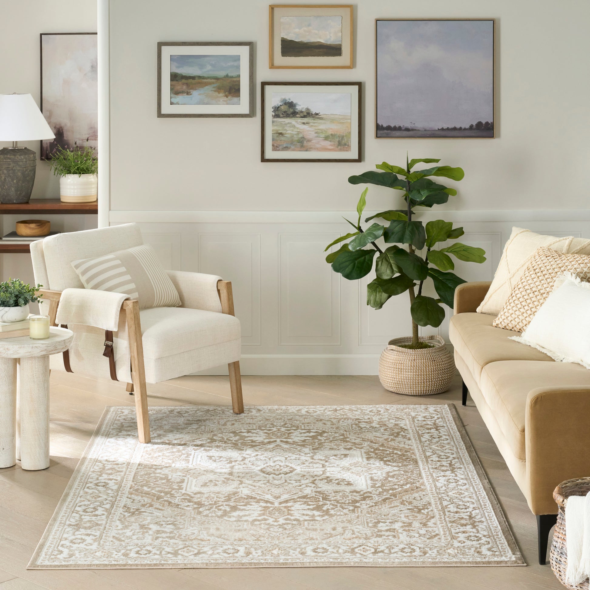 Cozy neutral living room with beige sofa, light wood armchair, ornate beige rug, fiddle-leaf fig, and framed landscape art.
