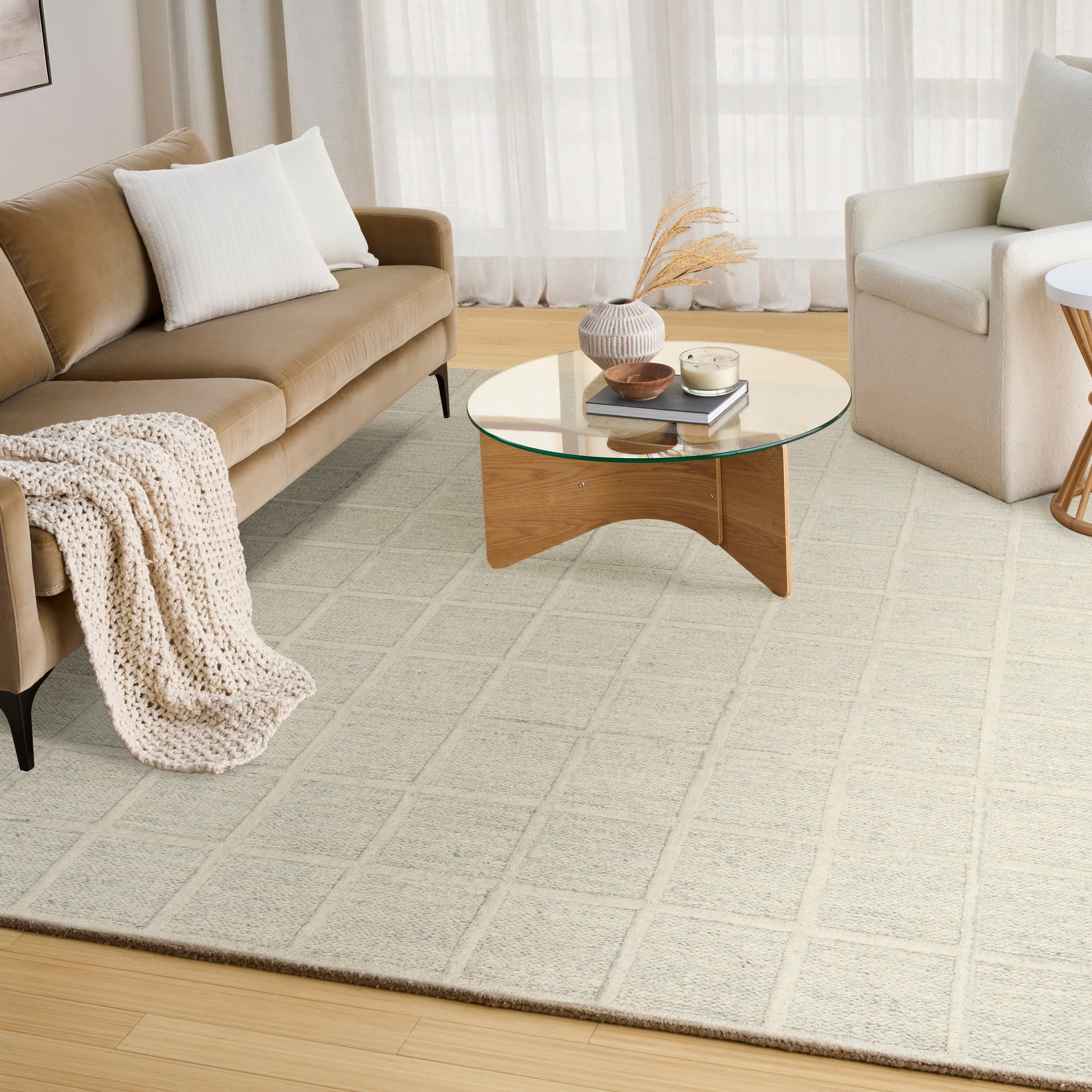 Cozy living room with tan sofa, white pillows, knit throw, round glass coffee table on light gray rug, cream armchair.