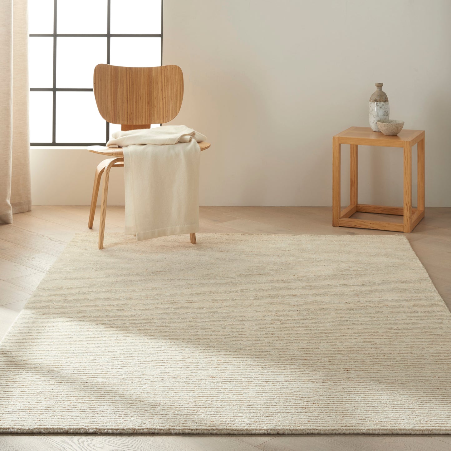 Natural wood chair with curved back, draped cream throw, beige woven rug, and a small wooden cube table with pottery.