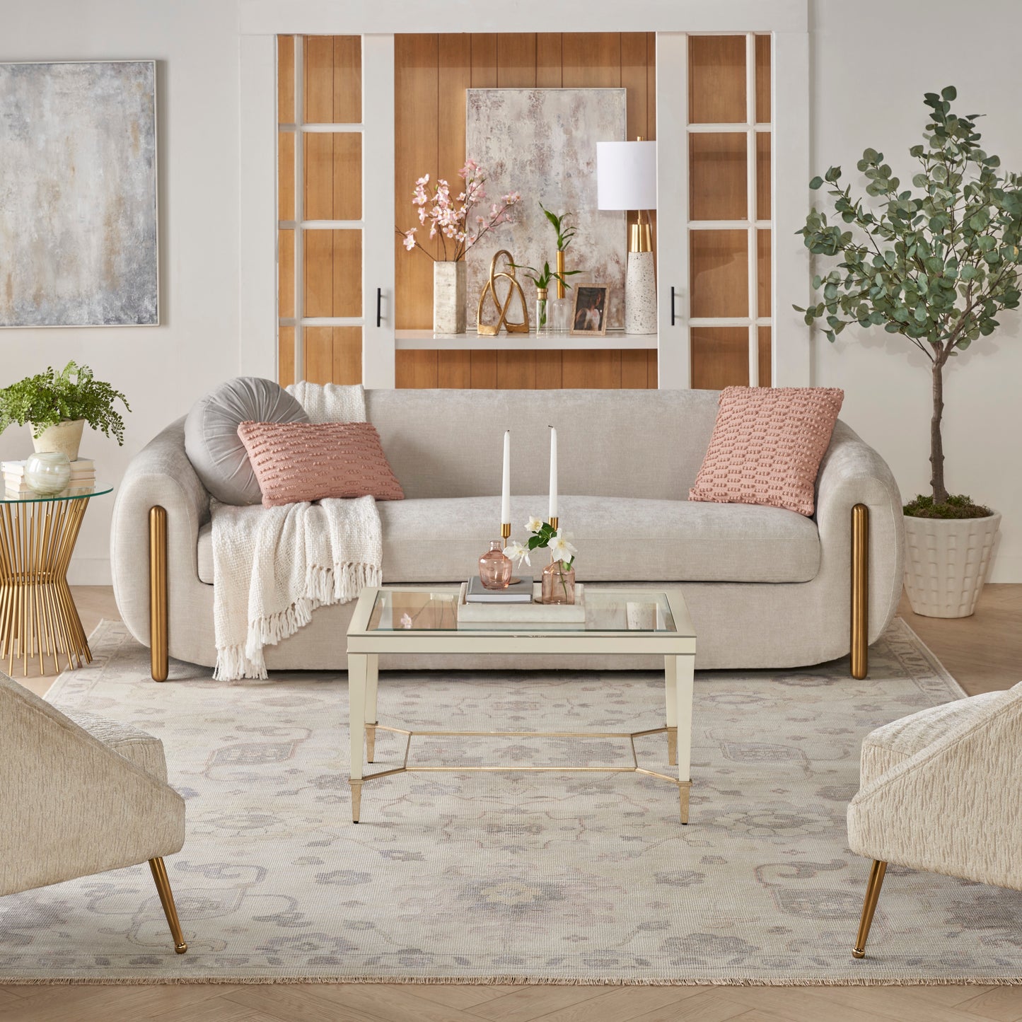 Neutral living room with beige sofa, pink cushions, glass coffee table, rug, and wood-framed display cabinet with plants.