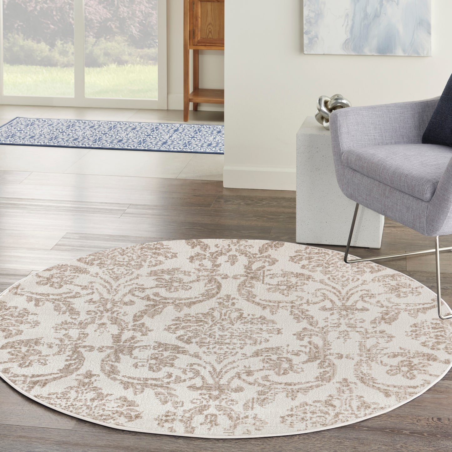 Round beige area rug with brown damask pattern on dark wood floor, in a living room with a gray chair and modern metal legs.