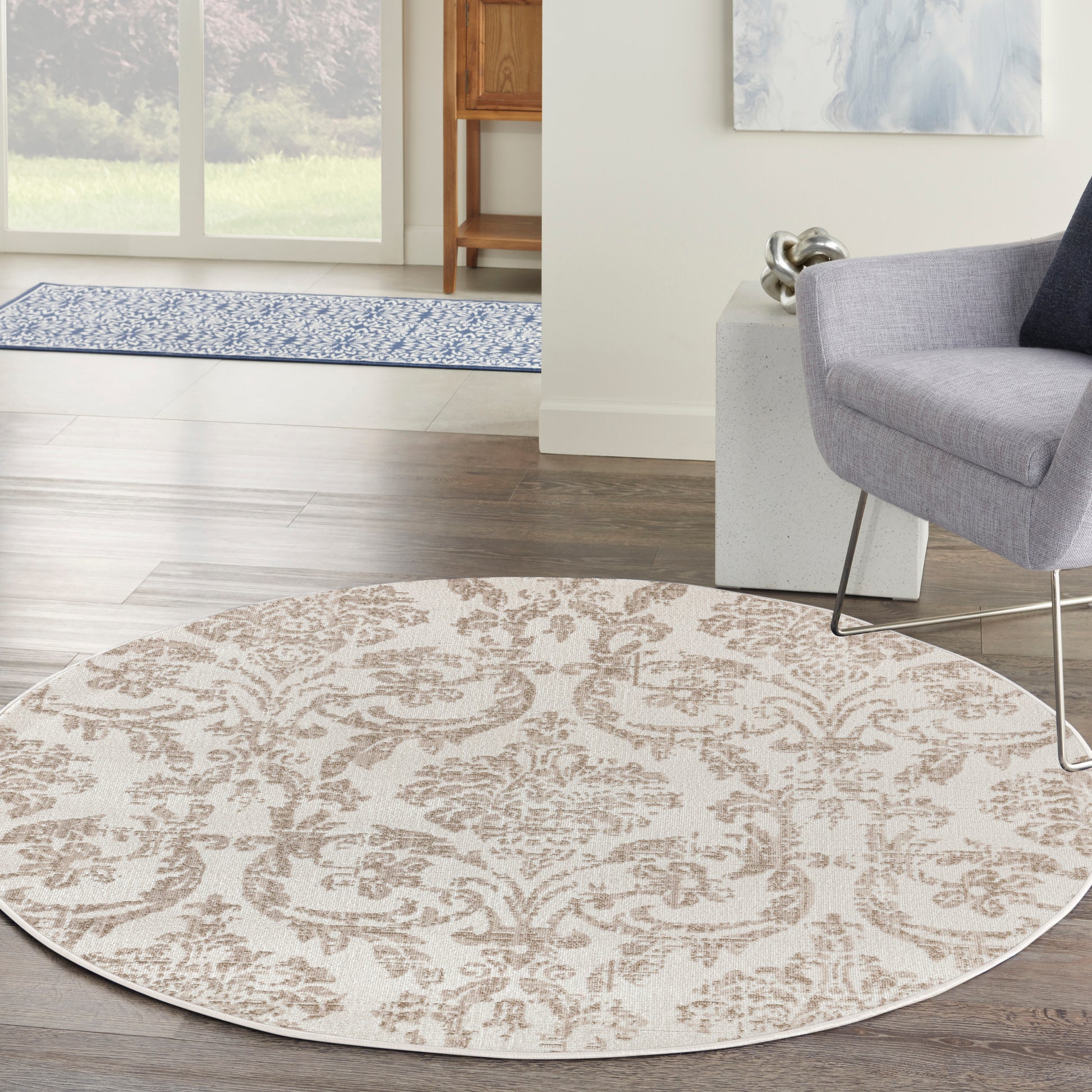Round beige area rug with brown damask pattern on dark wood floor, in a living room with a gray chair and modern metal legs.