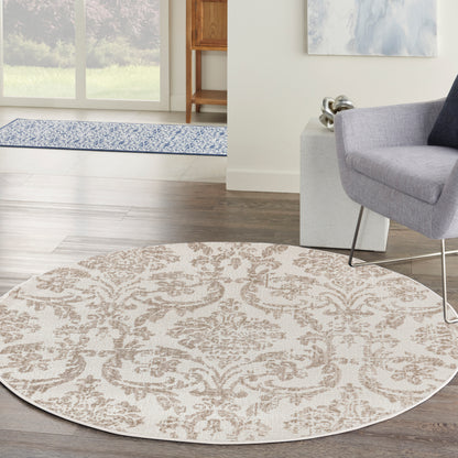 Round beige area rug with brown damask pattern on dark wood floor, in a living room with a gray chair and modern metal legs.