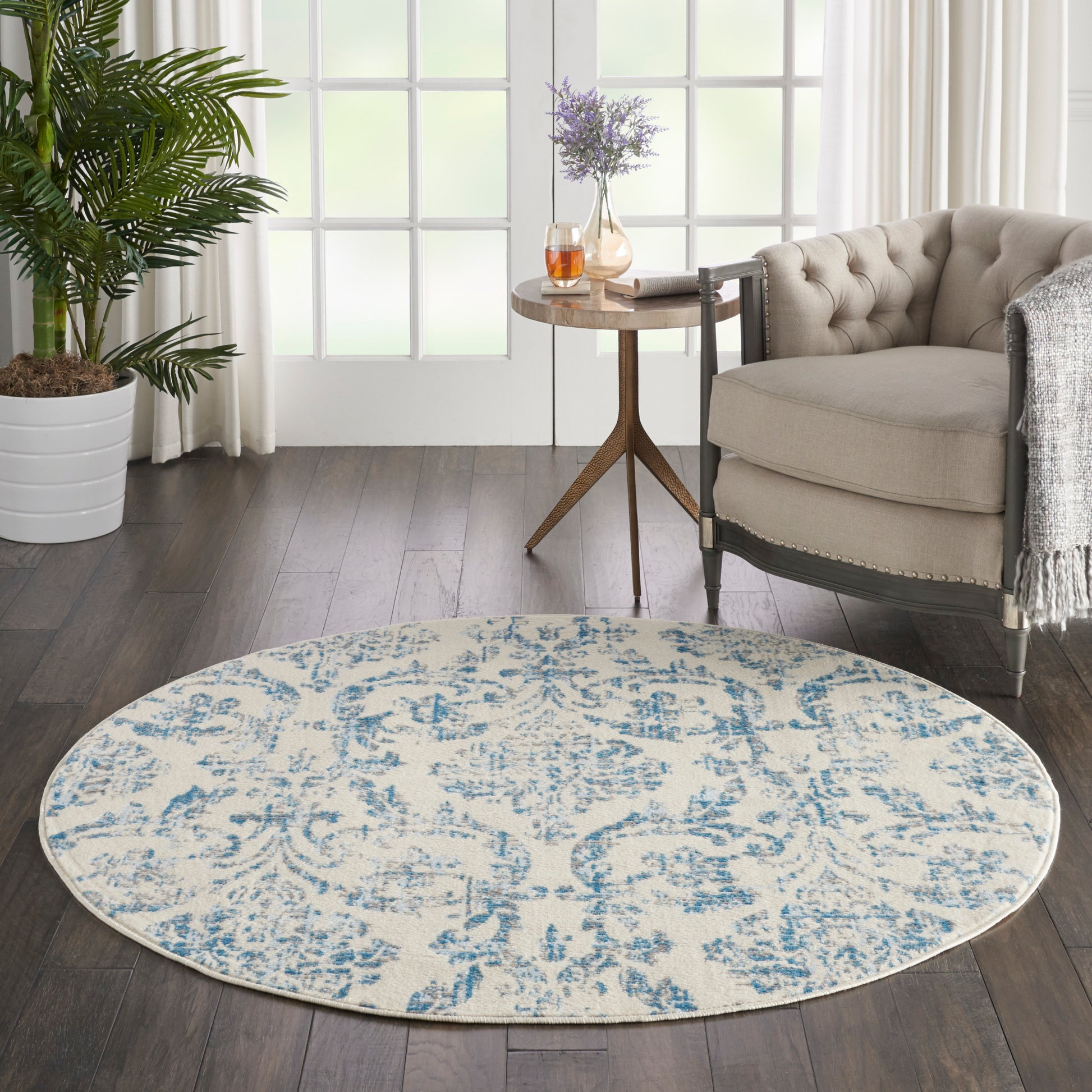 Round cream rug with blue damask pattern in a modern living room beside a tufted beige chair and a wooden side table.