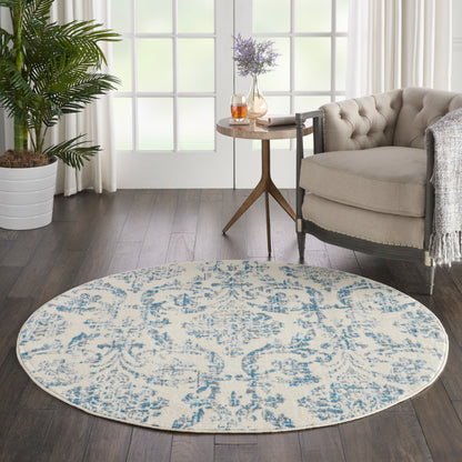 Round cream rug with blue damask pattern in a modern living room beside a tufted beige chair and a wooden side table.