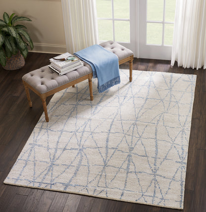 Cream flat-weave area rug with pale blue geometric lattice in a living room beside a tufted beige bench and blue throw.