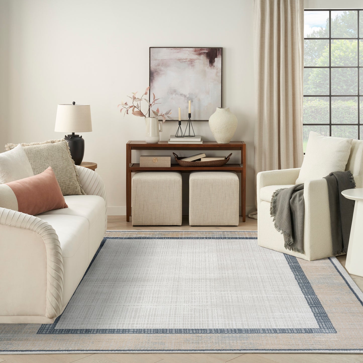 Neutral living room with beige sofa & cream armchair, wooden console, two ottomans, decorative vases, and a blue-bordered rug.