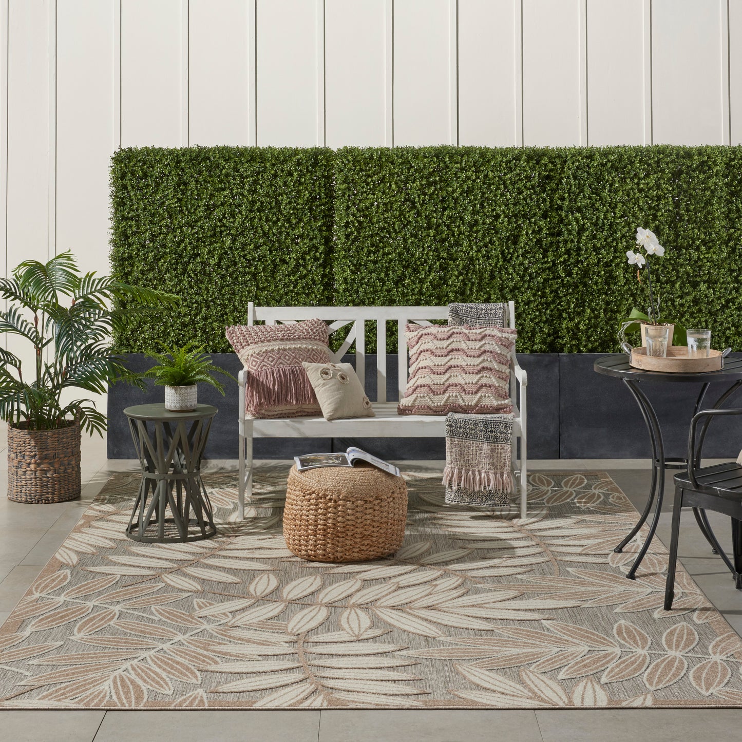Outdoor patio scene featuring a tropical-patterned rug (7' x 10') beneath a white bench, striped cushions, a round wicker ottoman, potted plants, and a green hedge backdrop.