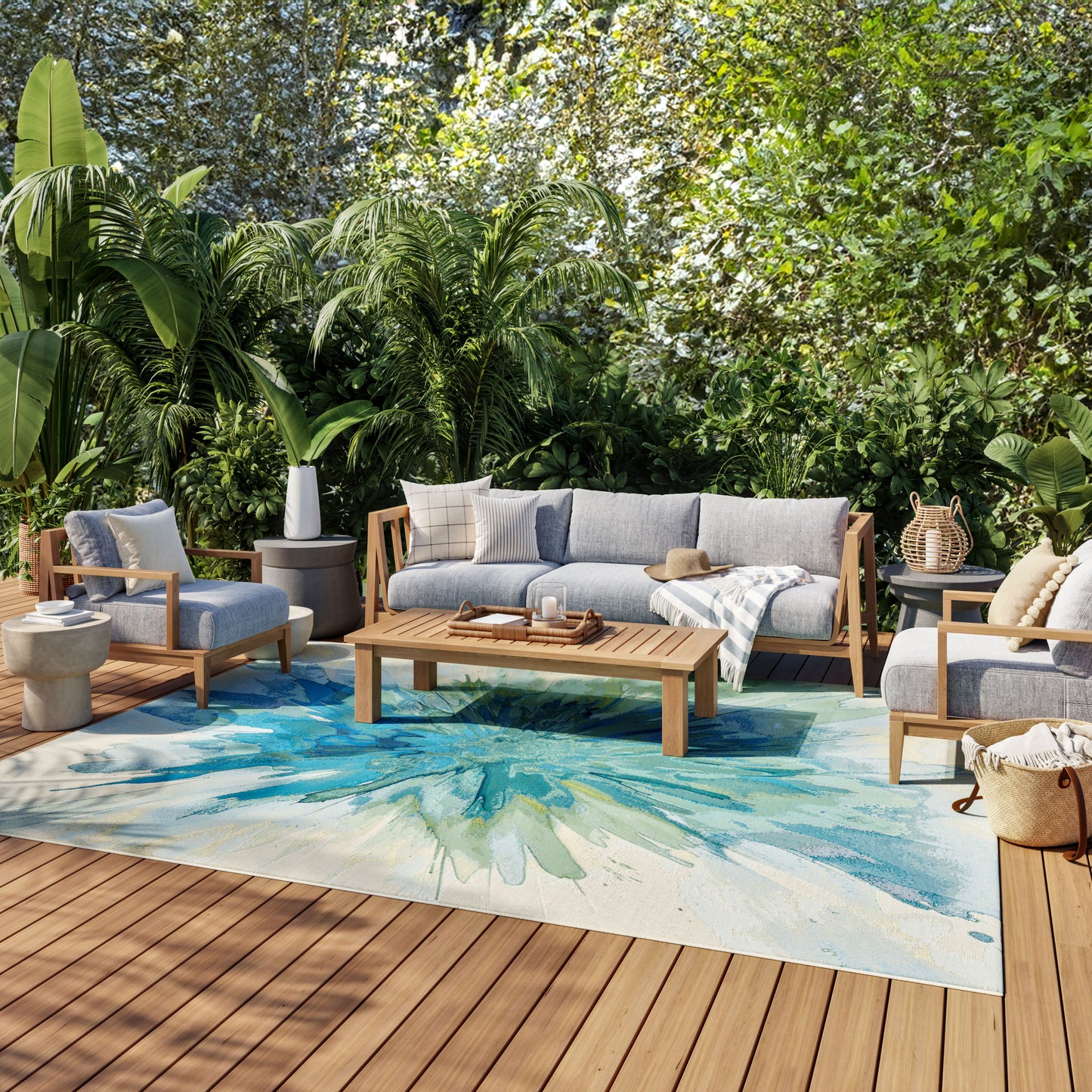 Outdoor patio lounge set with gray cushions on a wooden deck, tropical plants, and a blue-green abstract rug.