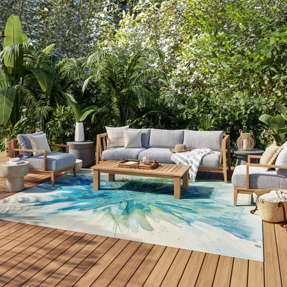 Outdoor patio lounge set with gray cushions on a wooden deck, tropical plants, and a blue-green abstract rug.