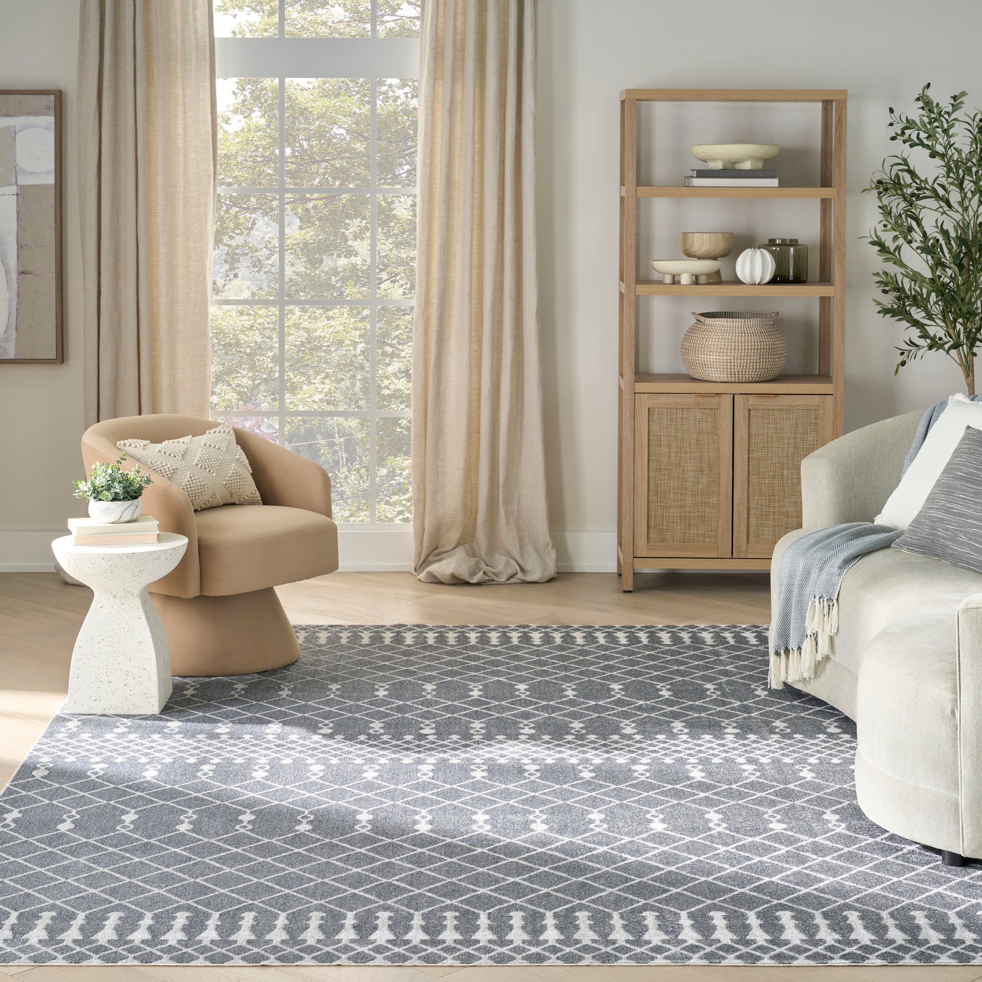 Sunlit living room with beige curved armchair, white round side table, gray geometric rug, wood shelving, sofa with blue throw.